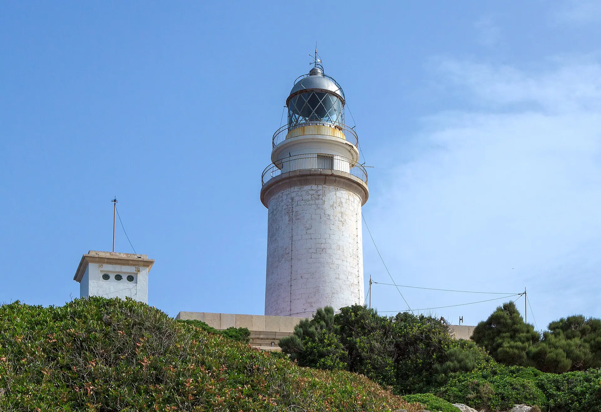 Weißer Leuchtturm am Cap de Formentor auf Mallorca vor blauem Himmel, im Vordergrund niedriges Buschwerk