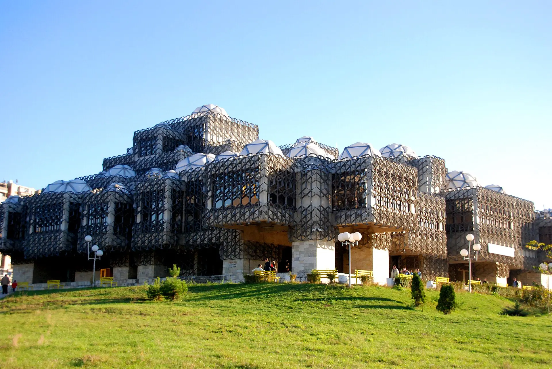 Nationalbibliothek in Pristina: gestaffelte Betonwürfel mit vielen weißen Kuppeln hinter einem dunklen Metallgitter, davor eine Wiese