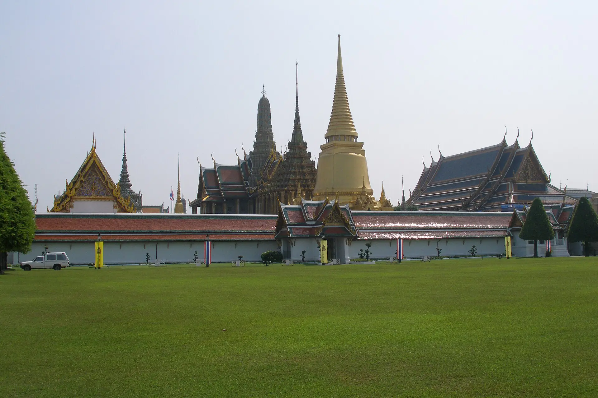 Tempelbezirk Wat Phra Kaeo hinter einer Rasenfläche: weiße Mauer, rot-grüne Dächer, goldener Chedi und Prangs