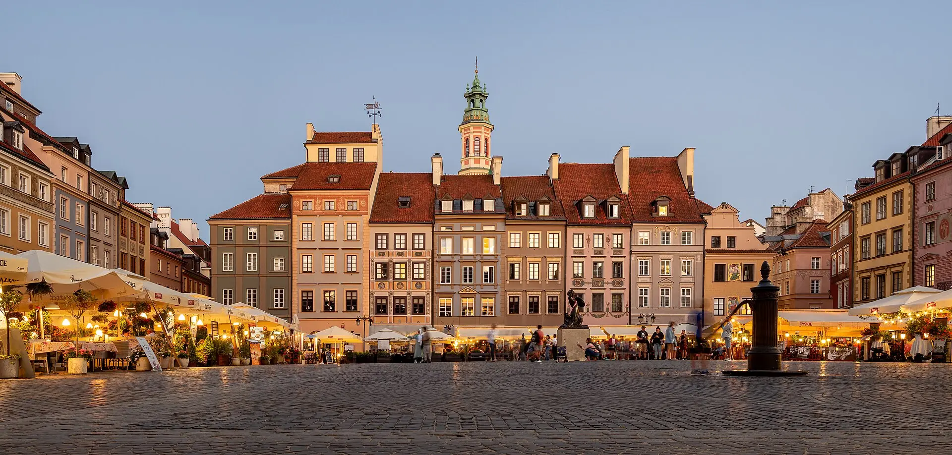 Warschauer Altstadtmarkt in der Dämmerung: farbige Bürgerhäuser mit roten Dächern, dahinter ein Turm mit grüner Haube, davor Café-Schirme