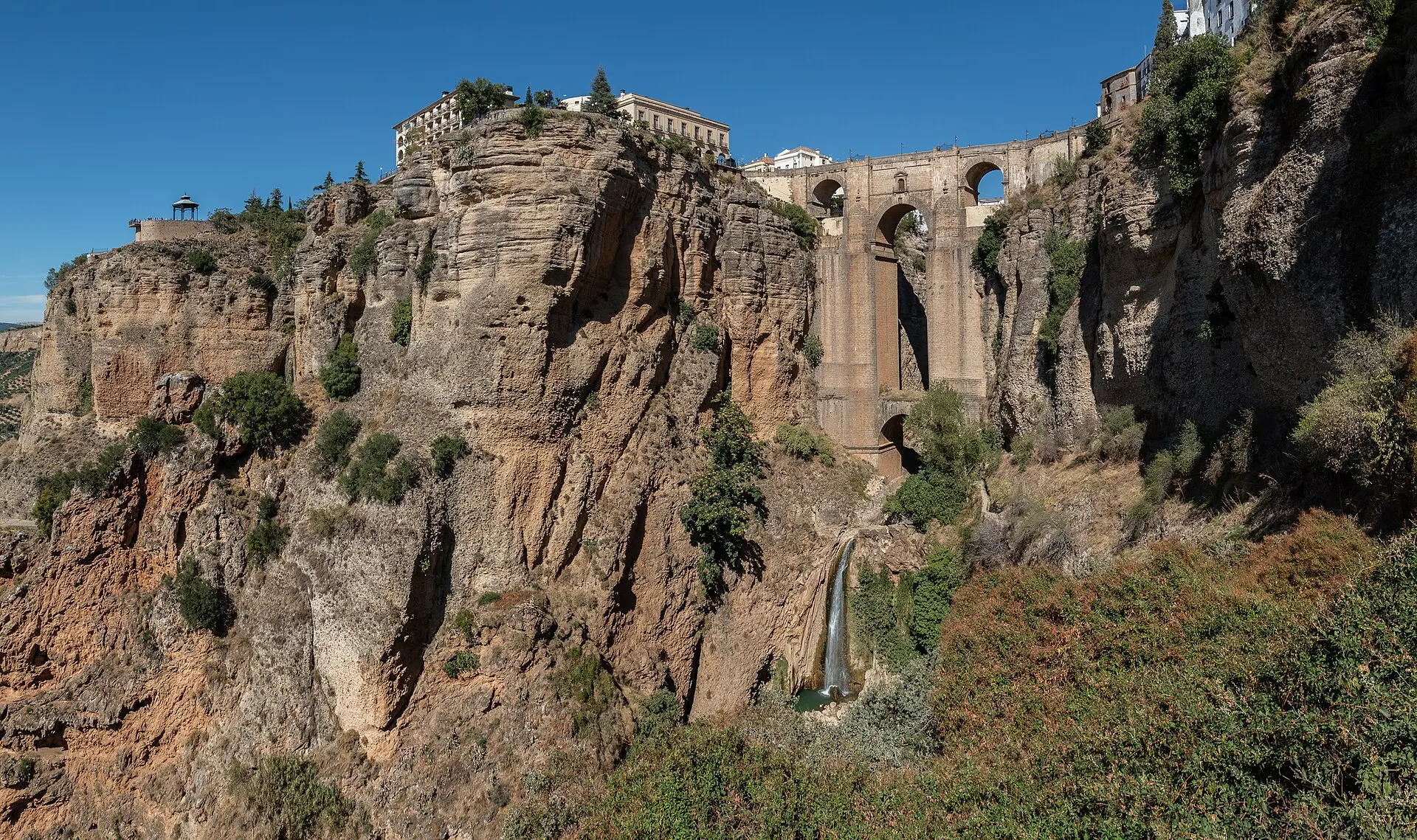 Puente Nuevo in Ronda zwischen zwei senkrechten Felswänden der Tajo-Schlucht, darunter ein Wasserfall, oben Häuser an der Kante