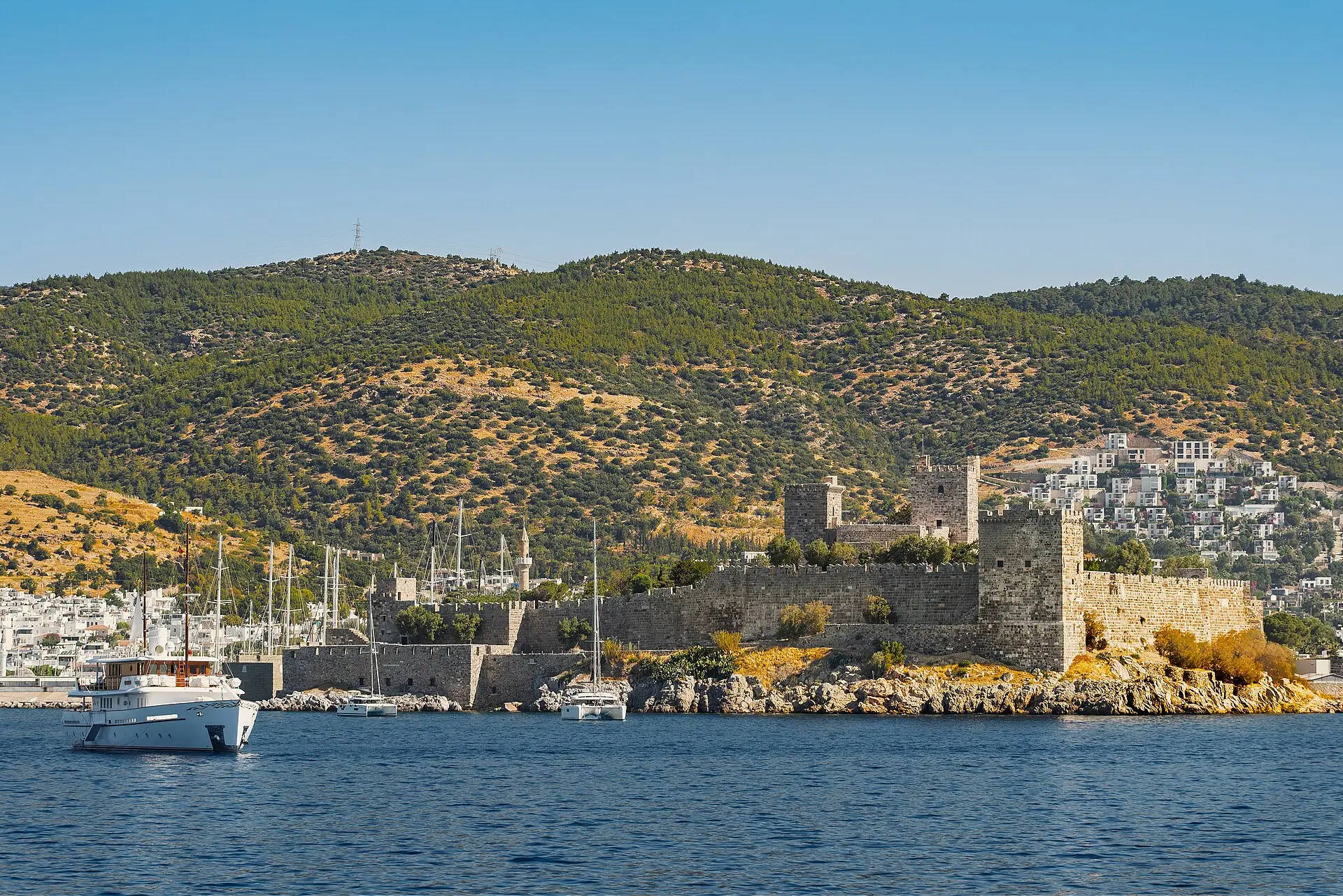 Burg von Bodrum mit grauen Mauern und eckigen Türmen auf einer felsigen Landzunge, links ein Jachthafen, dahinter Hügel mit weißen Häusern