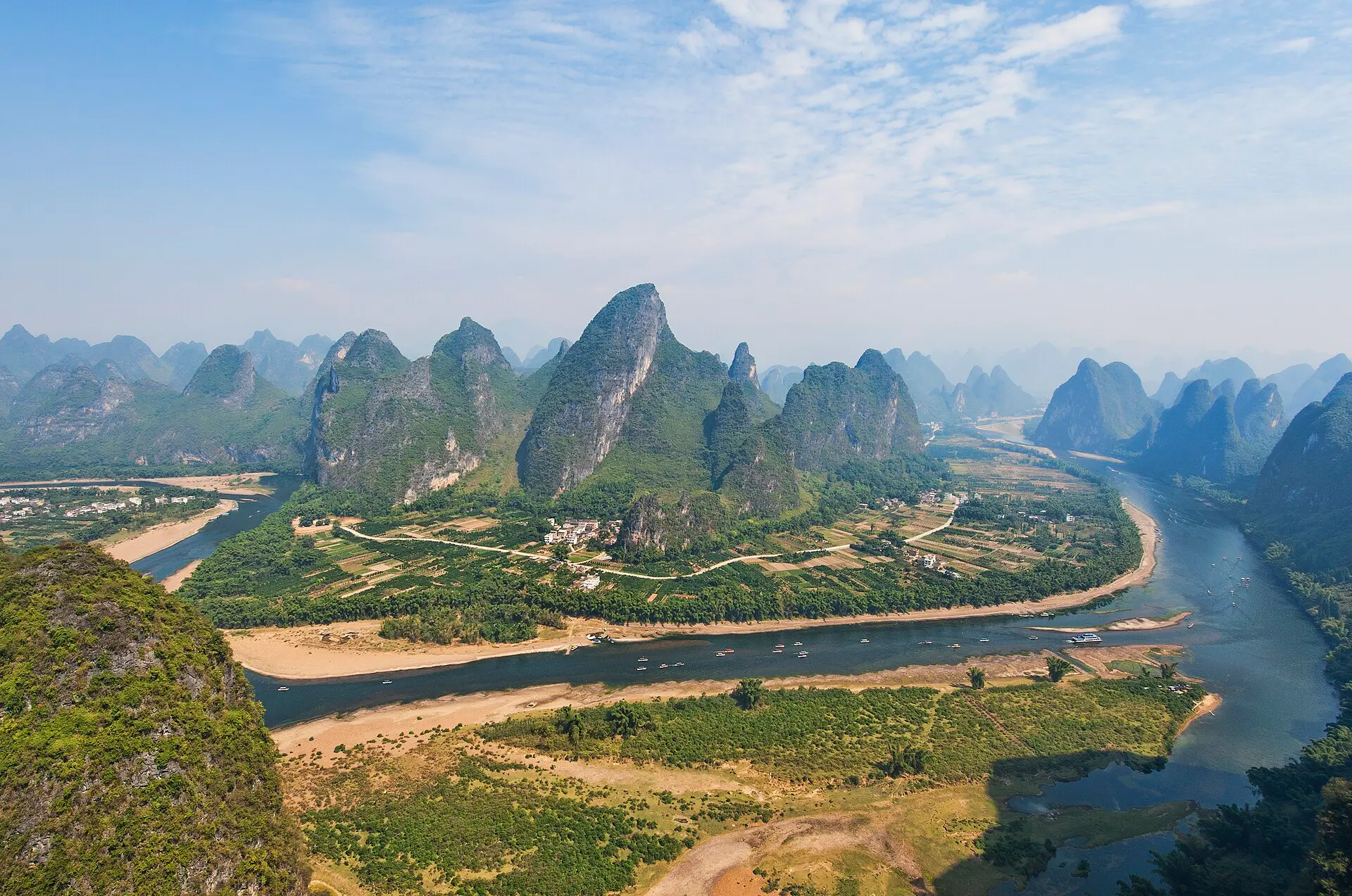Li-Fluss bei Yangshuo, von einem Hügel aus: Flussschleife zwischen Feldern und einem Dorf, kleine Boote, dahinter Karstkegel im Dunst