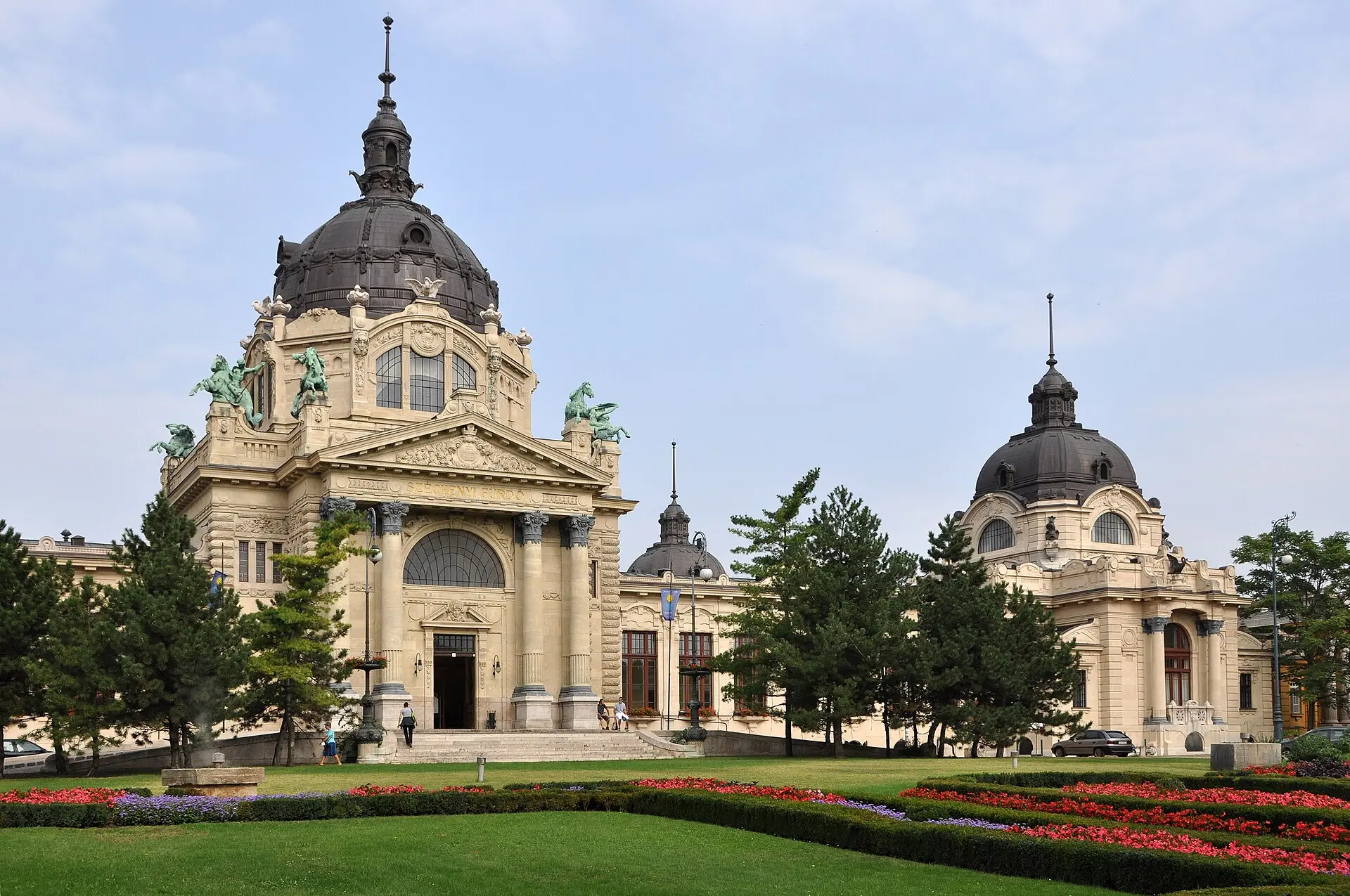 Hauptgebäude des Széchenyi-Bads mit großer dunkler Kuppel und Figurengruppen, davor Rasen mit roten Blumenbeeten