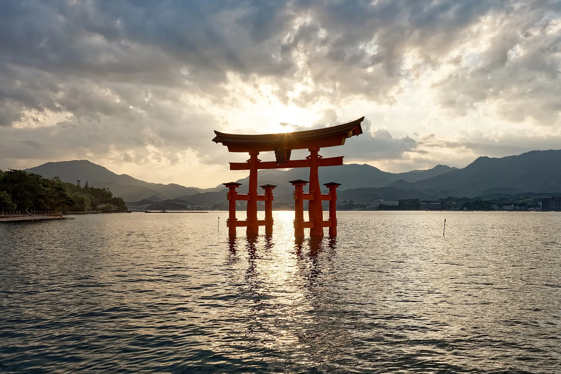 Rotes Torii des Itsukushima-Schreins steht bei Flut im Wasser, Gegenlicht der tief stehenden Sonne, dahinter Bergketten