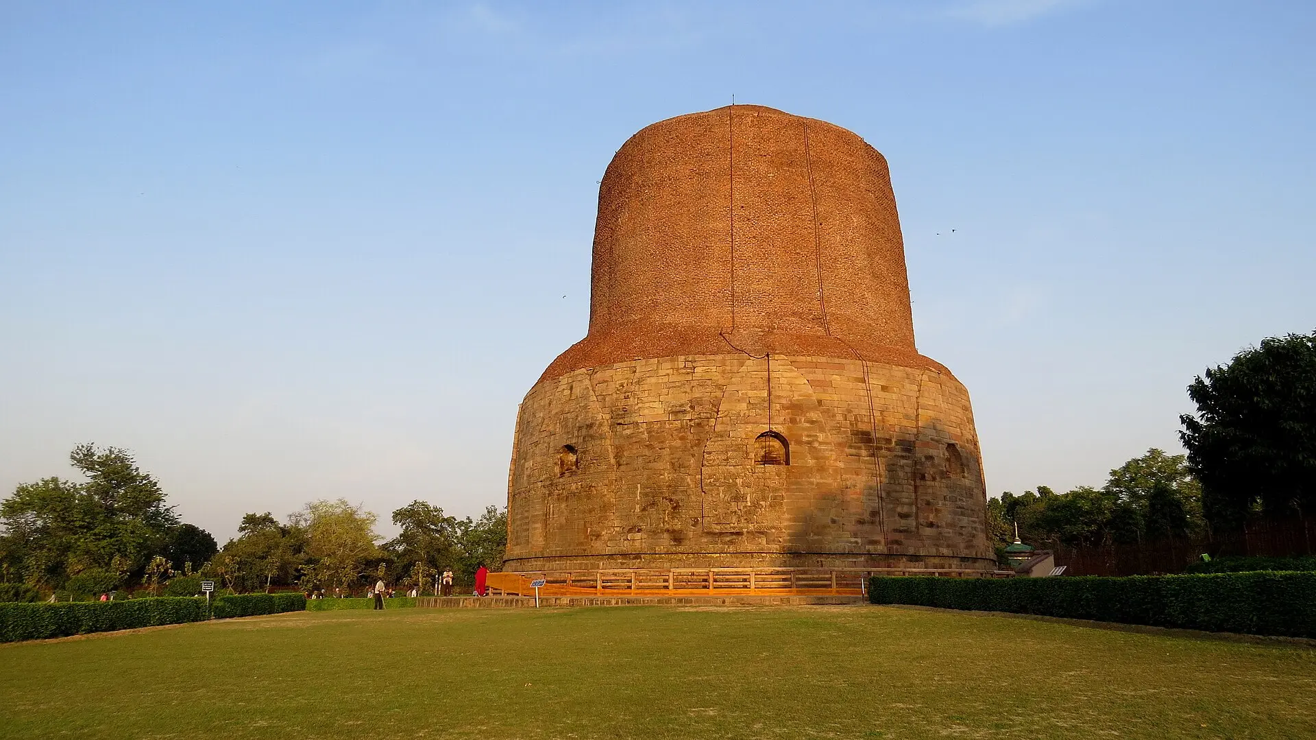 Runder Dhamek-Stupa in Sarnath, unten helle Steinverkleidung, oben rotbraunes Mauerwerk, auf Rasen mit Hecken im Abendlicht