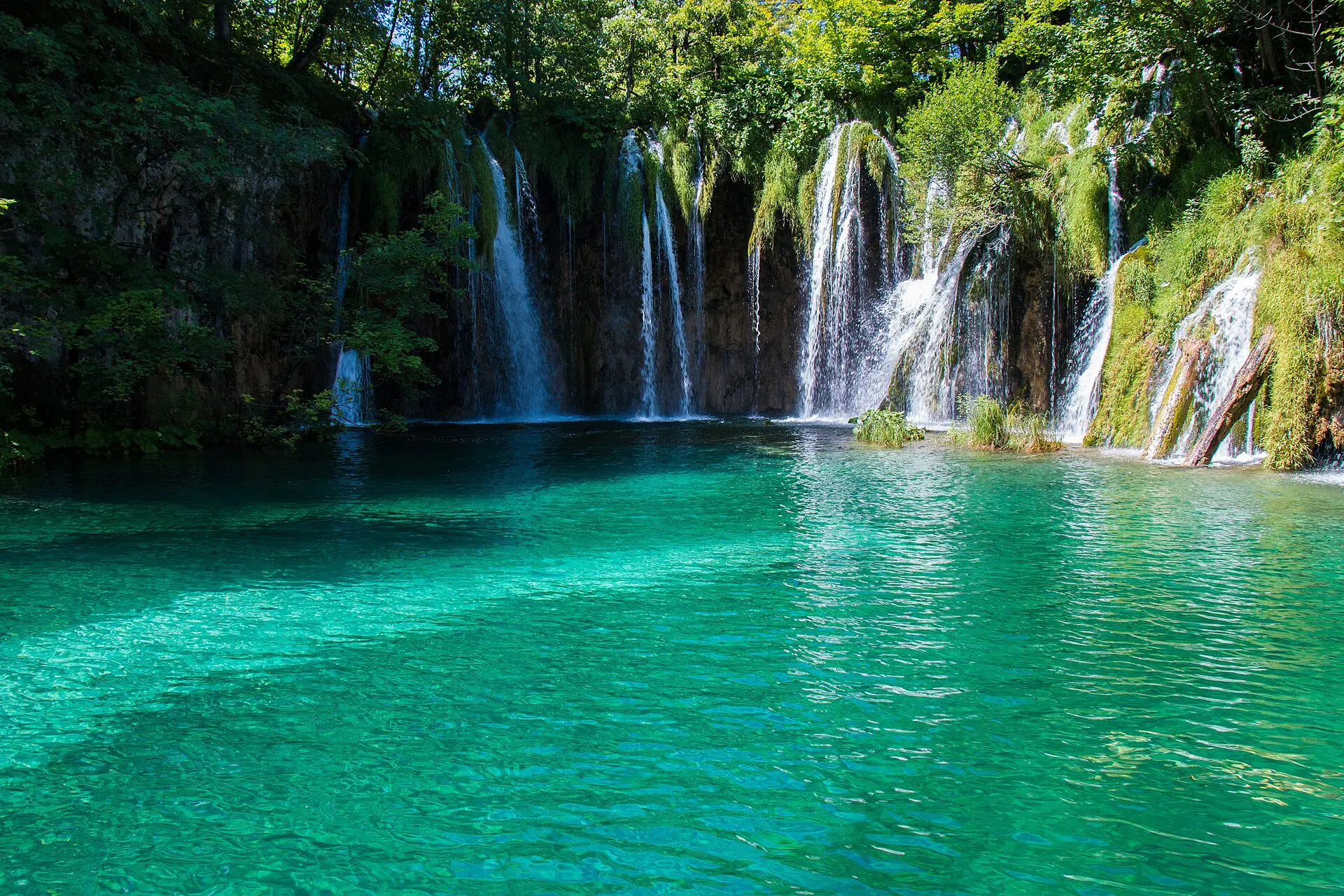 Mehrere schmale Wasserfälle fallen über eine bemooste Felswand in einen türkisgrünen See im Nationalpark Plitvicer Seen