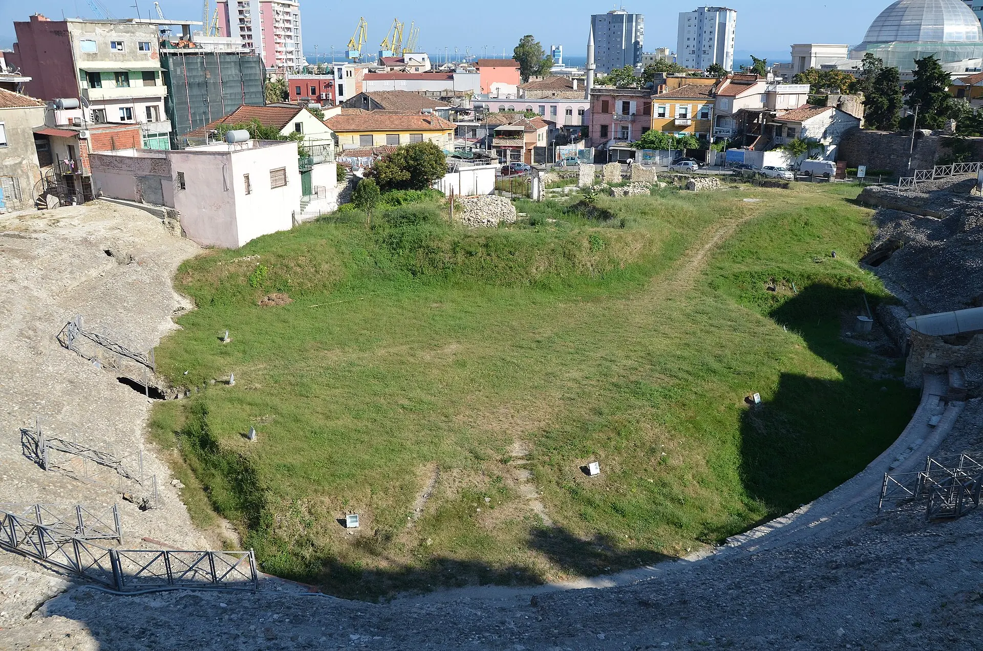 Blick von den oberen Sitzreihen auf die begrünte Arena des Amphitheaters von Durrës, ringsum Wohnhäuser, dahinter Hafenkräne