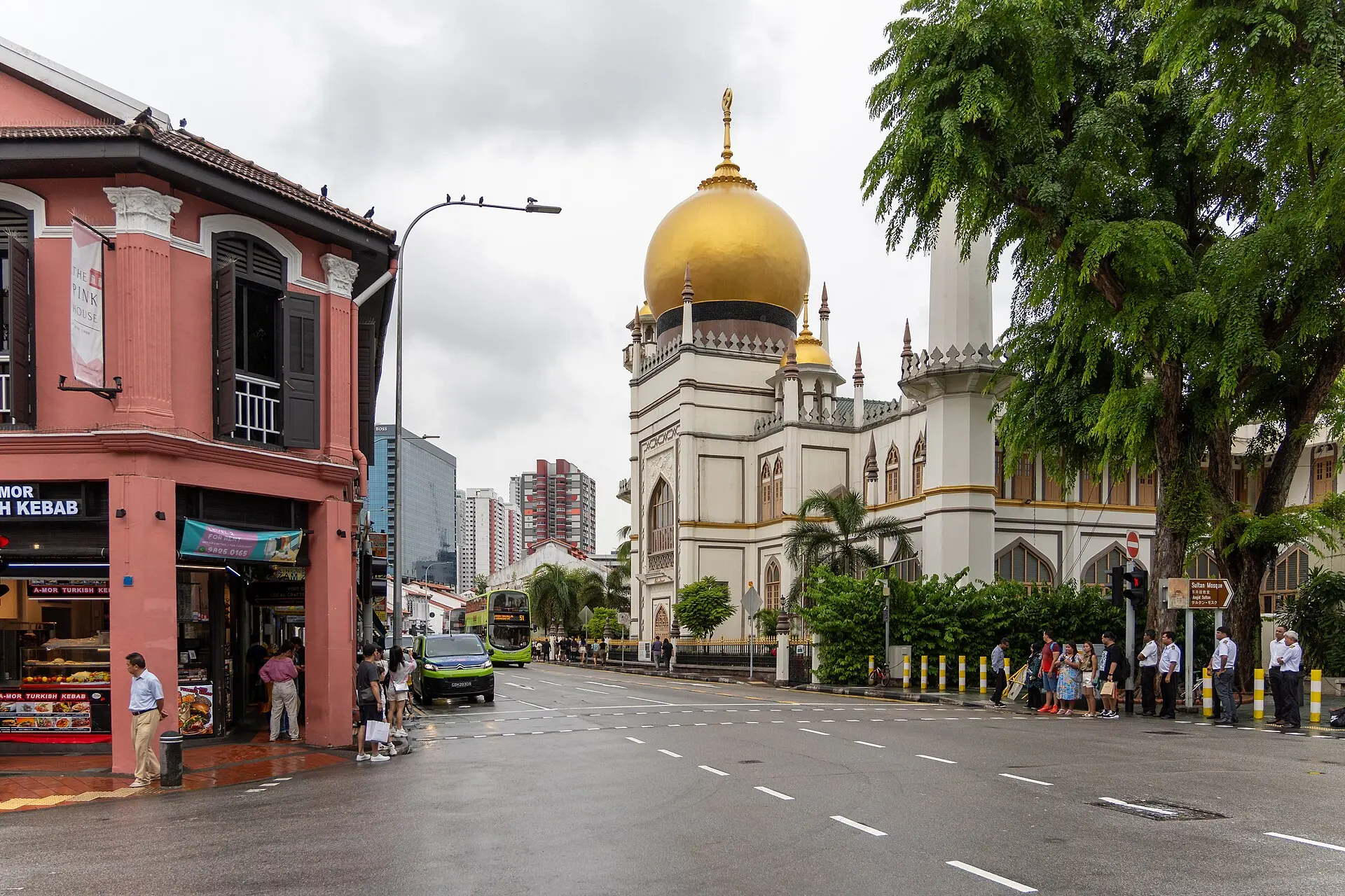Sultan-Moschee mit goldener Kuppel und Minarett, links ein rosafarbenes Eckhaus, vorn eine nasse Straße, dahinter ein grüner Doppeldeckerbus