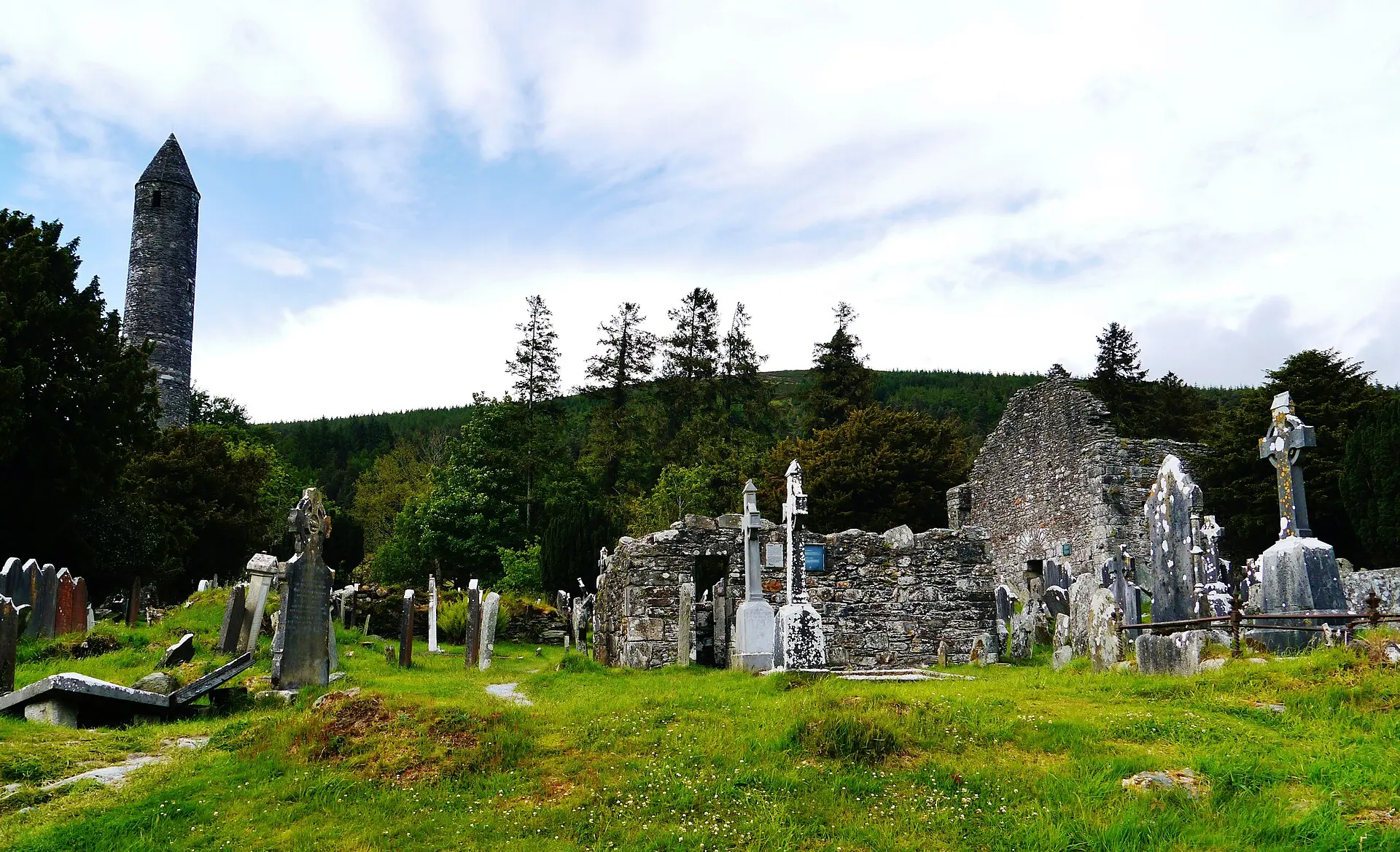 Der schlanke Rundturm von Glendalough mit Kegeldach, davor Grabsteine und Keltenkreuze, rechts eine Kirchenruine aus Bruchstein
