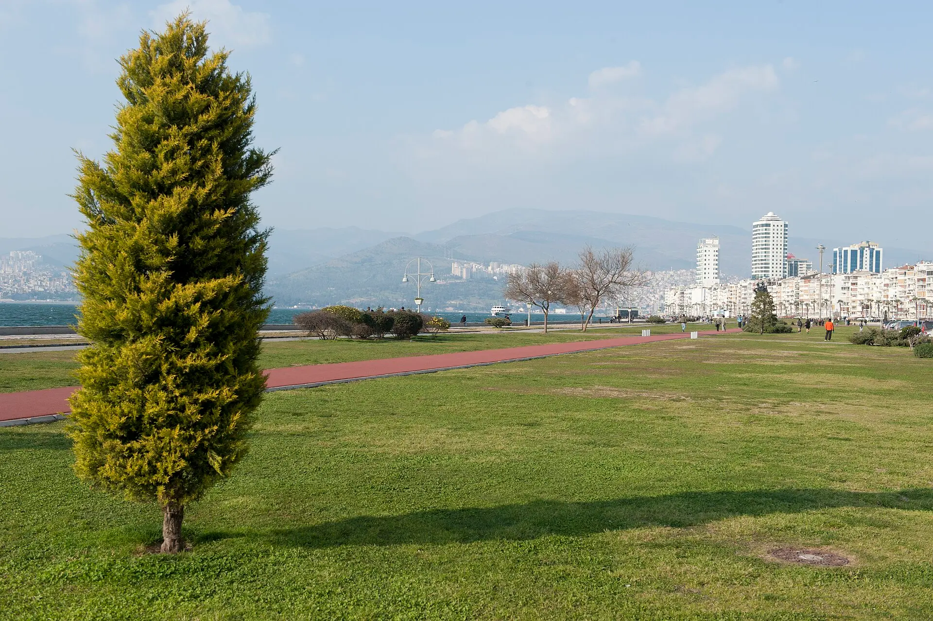 Grünfläche am Kordon in Izmir mit Zypresse und rotem Weg, dahinter die Uferlinie der Bucht, Hochhäuser und Berge