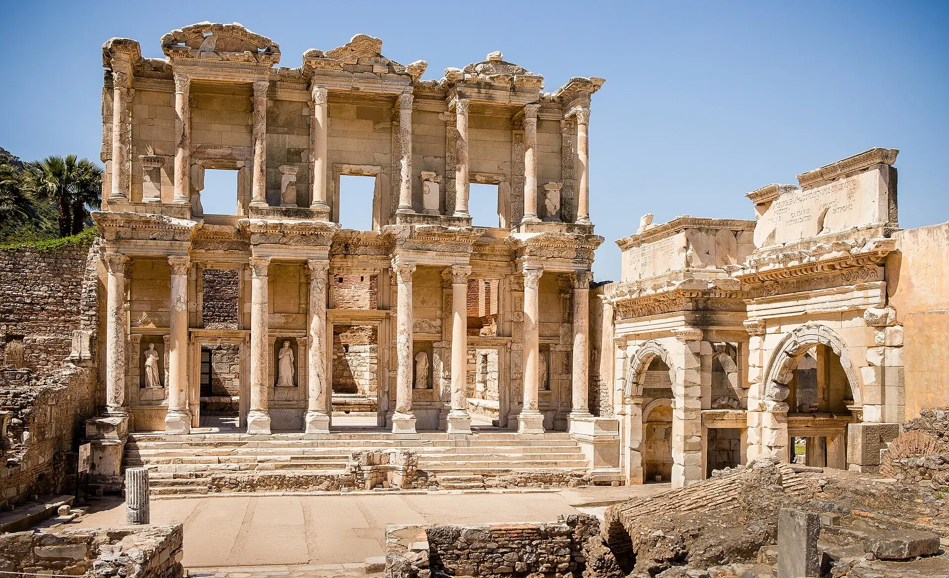 Fassade der Celsus-Bibliothek in Ephesos unter klarem blauem Himmel, rechts ein antikes Tor, vorn Mauerreste und eine Säule