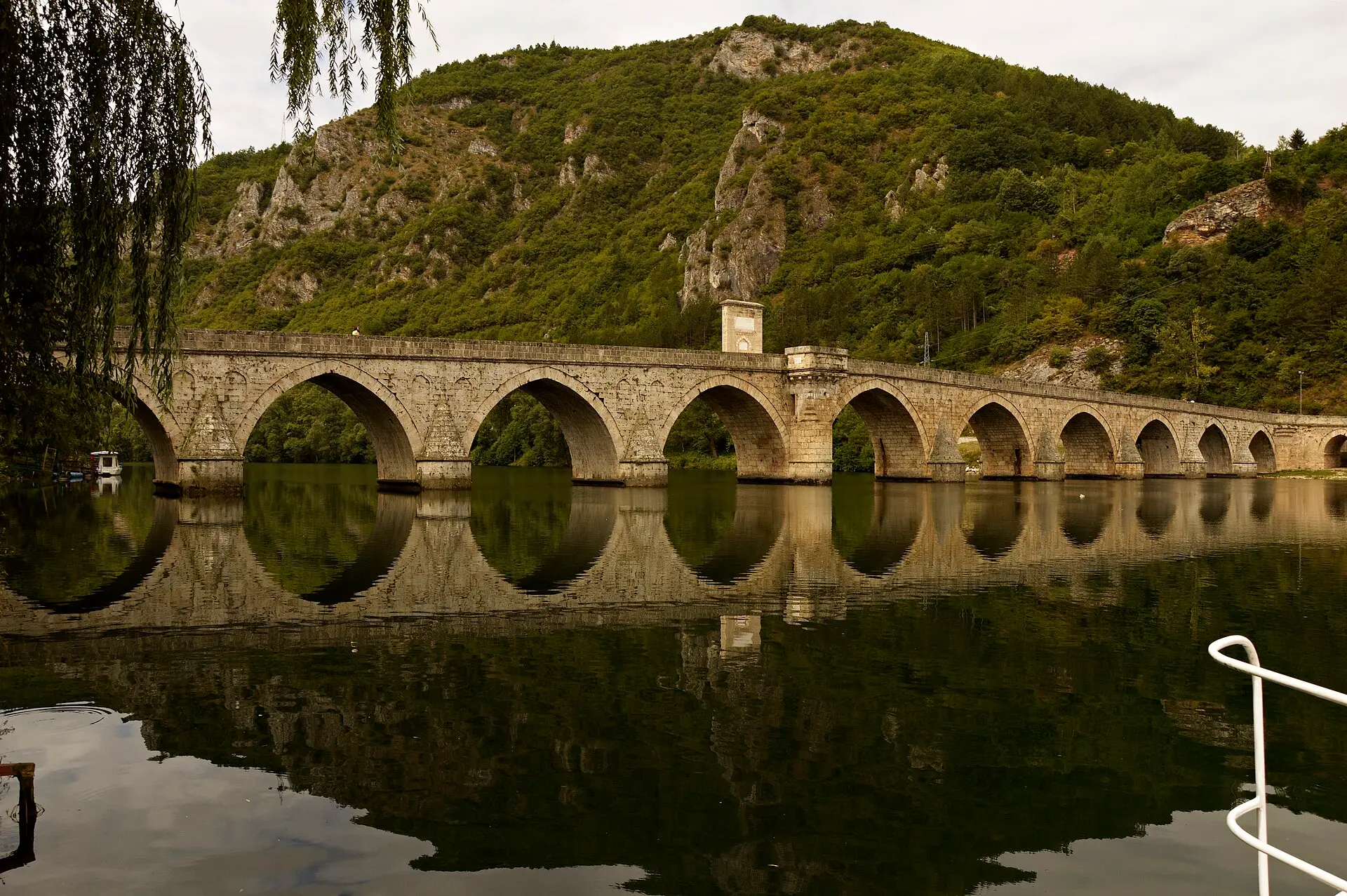 Mehmed-Paša-Sokolović-Brücke in Višegrad: lange Steinbrücke mit vielen Bögen über der ruhigen Drina, dahinter ein bewaldeter Berg