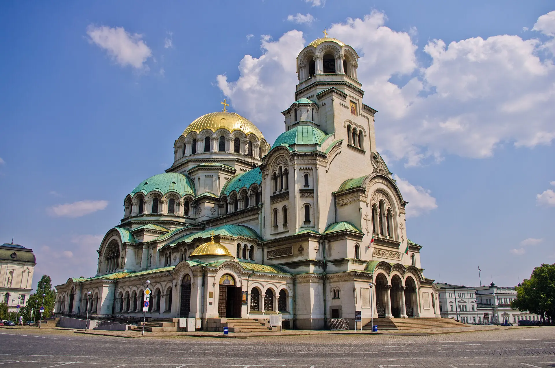 Alexander-Newski-Kathedrale in Sofia mit Glockenturm, goldenen und grünen Kuppeln, davor ein leerer gepflasterter Platz unter blauem Himmel