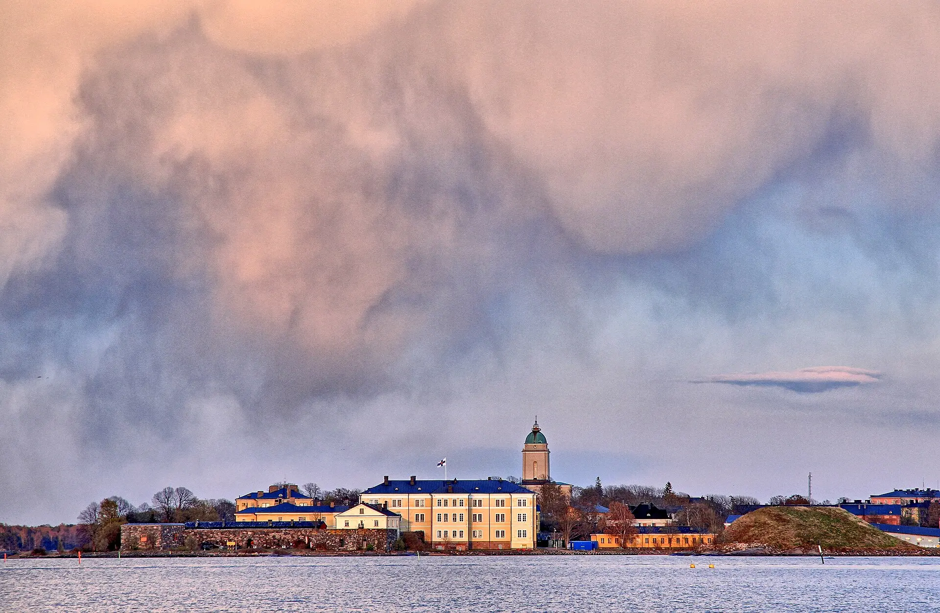 Suomenlinna vom Wasser aus: gelbe Häuser mit blauen Dächern, eine Steinmauer und ein Kirchturm mit grüner Kuppel im Abendlicht