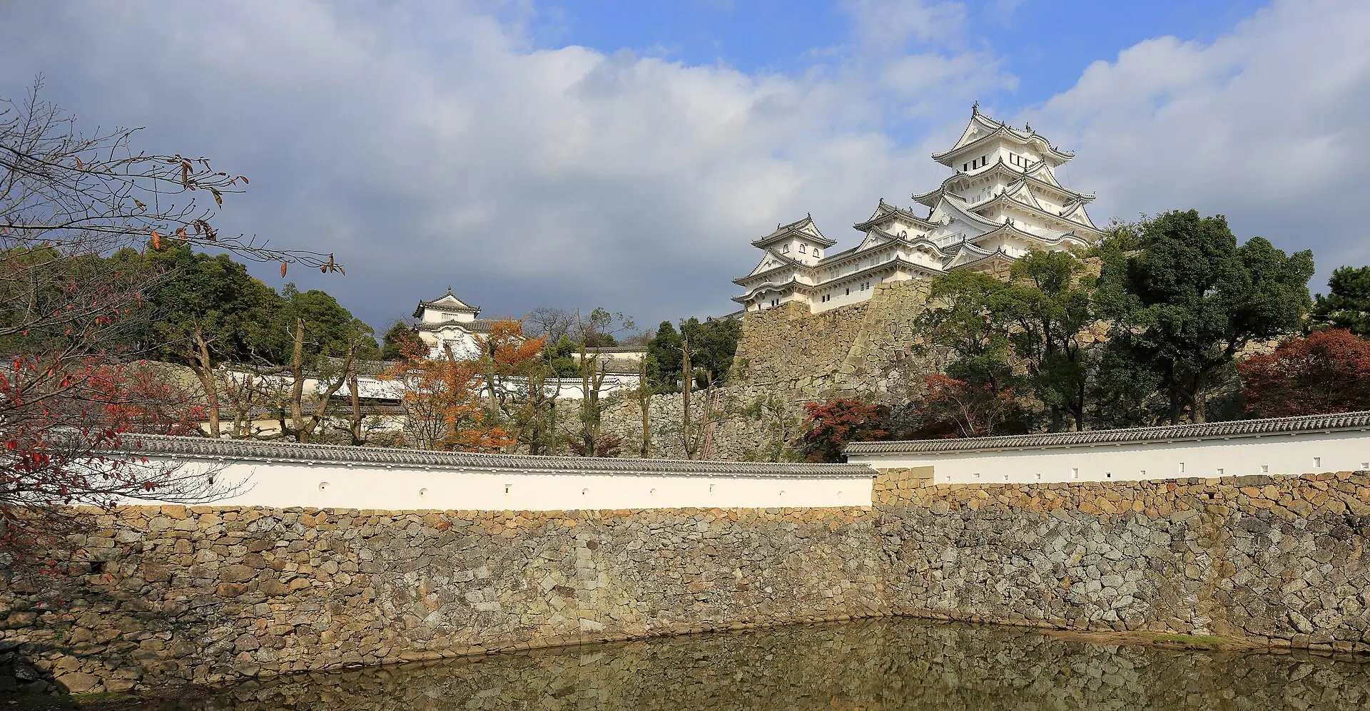 Weißer Hauptturm der Burg Himeji auf hohen Steinmauern, davor Burggraben, Putzmauer mit Ziegeldach und rot gefärbte Bäume
