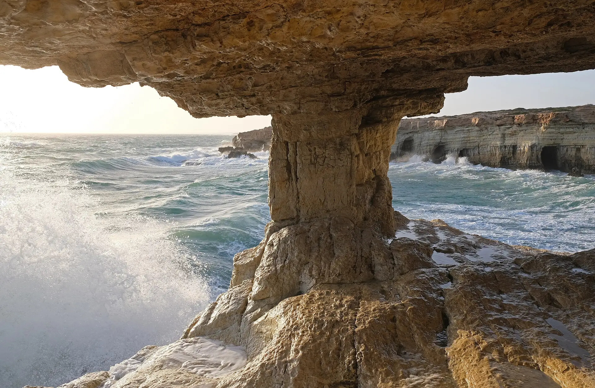 Blick durch ein Felsdach auf einer Säule auf das türkisgrüne Meer mit Brandung, rechts eine helle Steilküste mit Höhlen