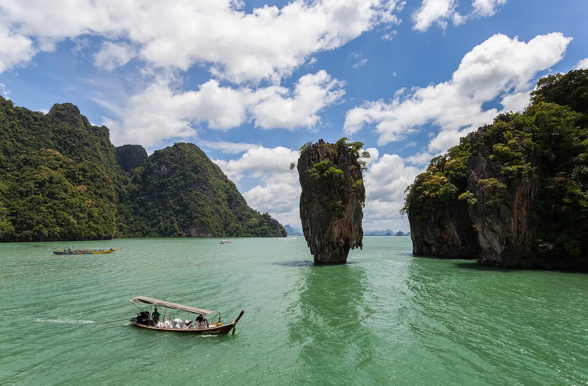 Kalksteinnadel Ko Tapu im grünen Wasser der Bucht von Phang-nga, daneben bewaldete Karstfelsen, vorn ein Longtailboot