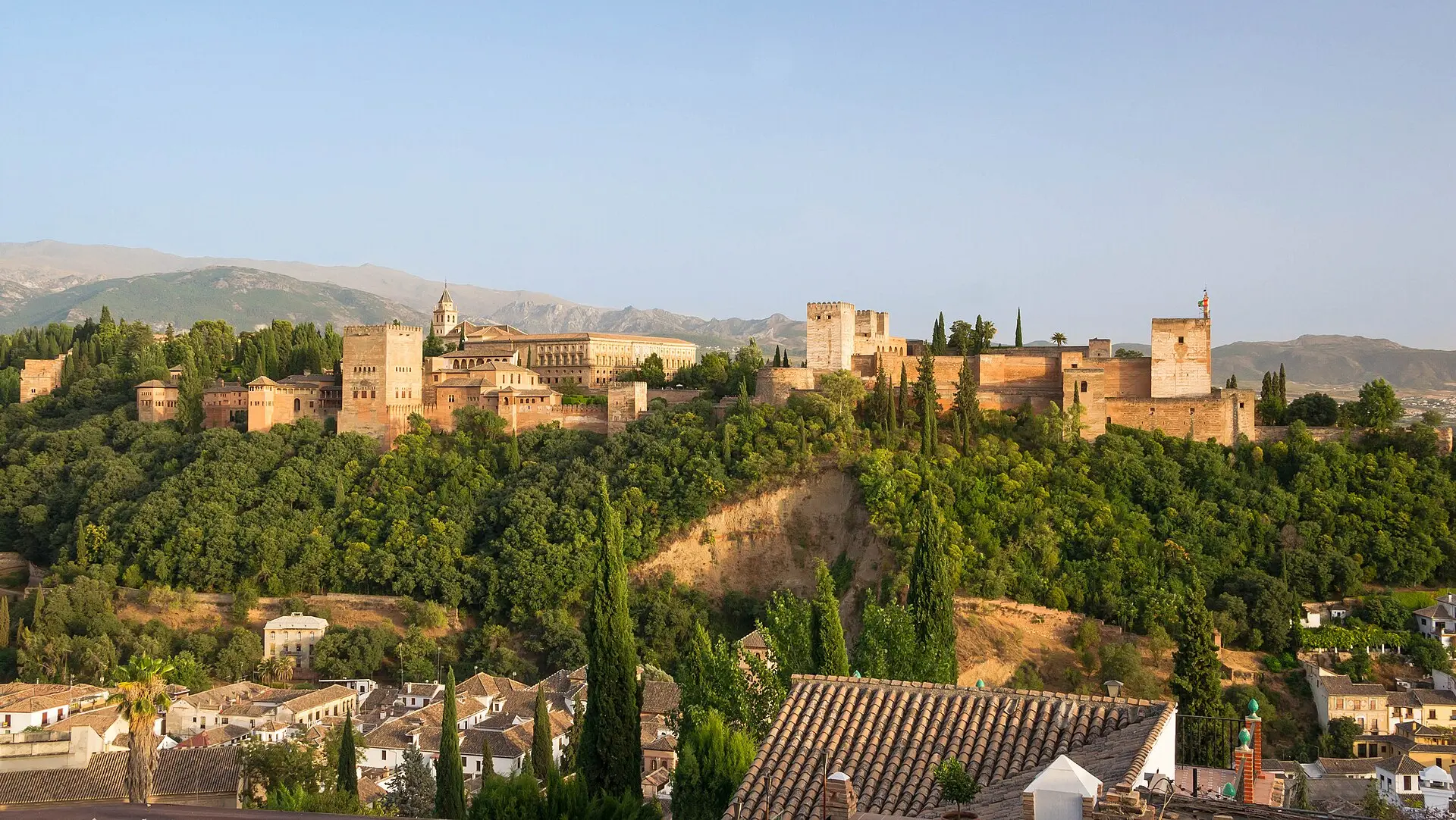 Alhambra in Granada im Abendlicht: Mauern und Türme auf einem bewaldeten Hügel, davor Ziegeldächer und Zypressen, dahinter Berge