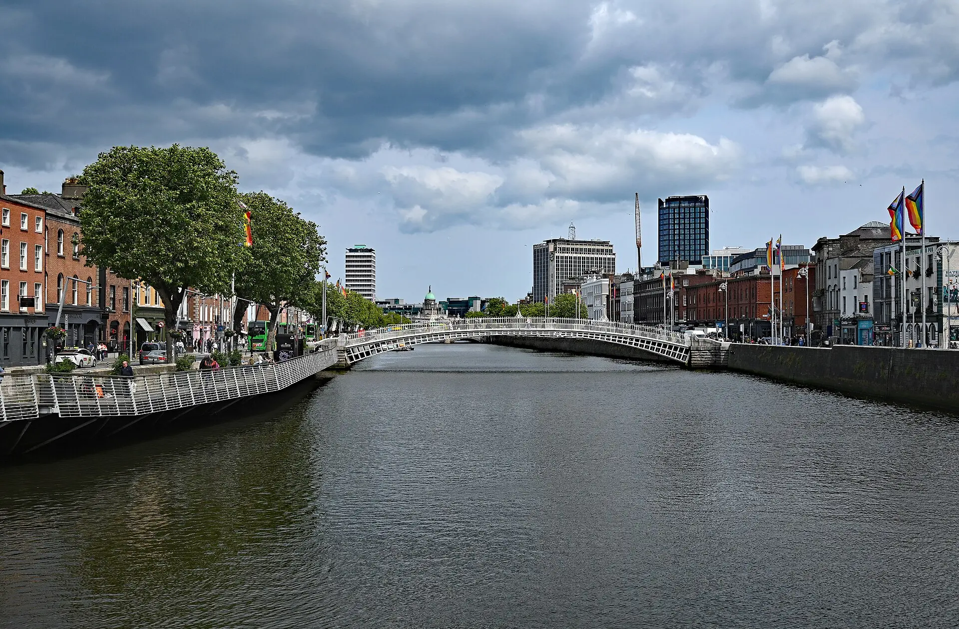Die Liffey zwischen Kaimauern, in der Bildmitte die weiße Bogenbrücke Ha’penny Bridge, dahinter Bürotürme unter dunklen Wolken