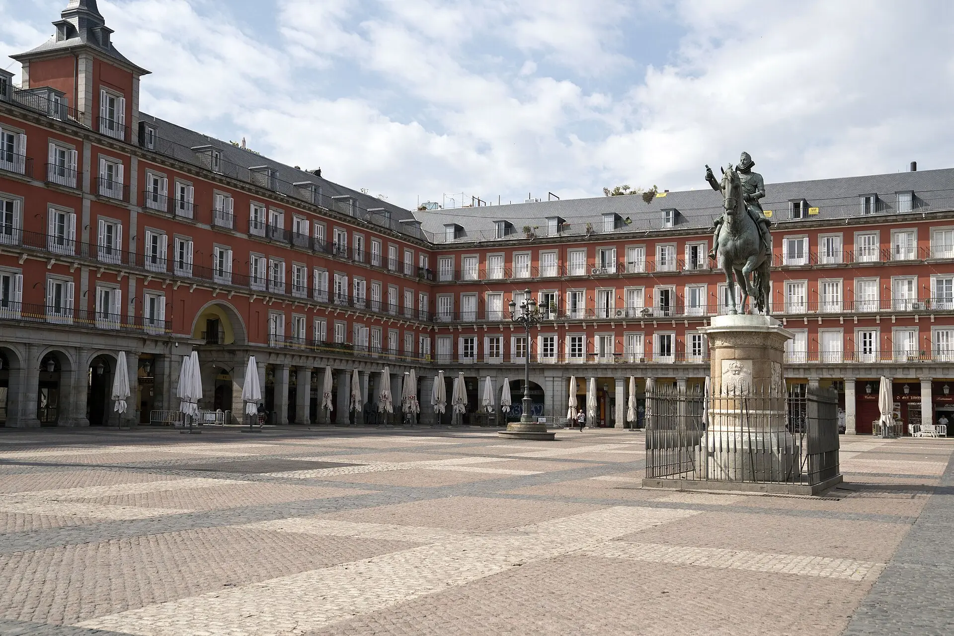 Plaza Mayor in Madrid bei teils bewölktem Himmel: rote Fassaden mit Balkonen, zugeklappte Sonnenschirme, rechts das Reiterstandbild Philipps III.