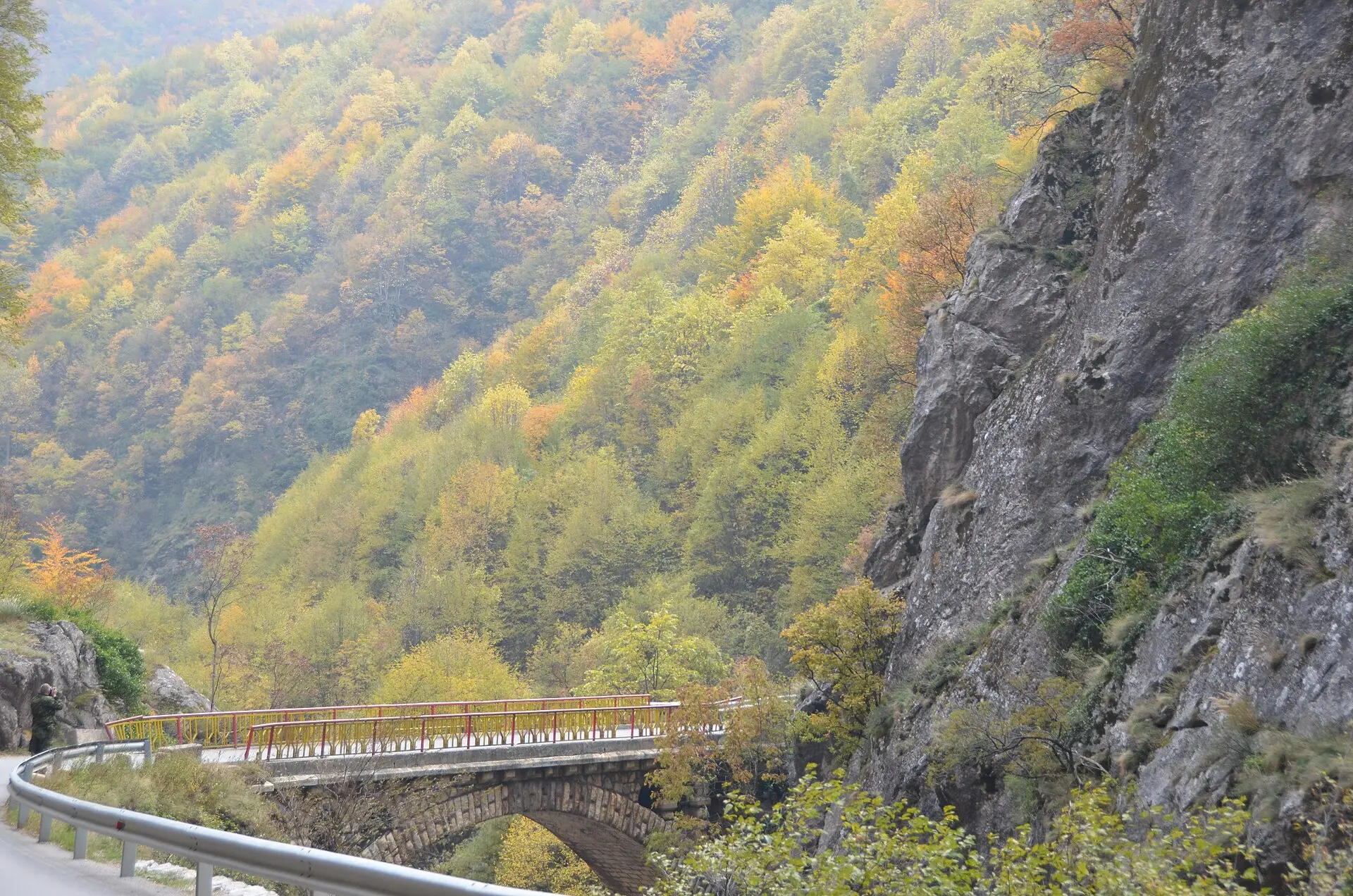 Steinerne Bogenbrücke mit rotem Geländer in der Rugova-Schlucht, daneben eine graue Felswand und Herbstwald