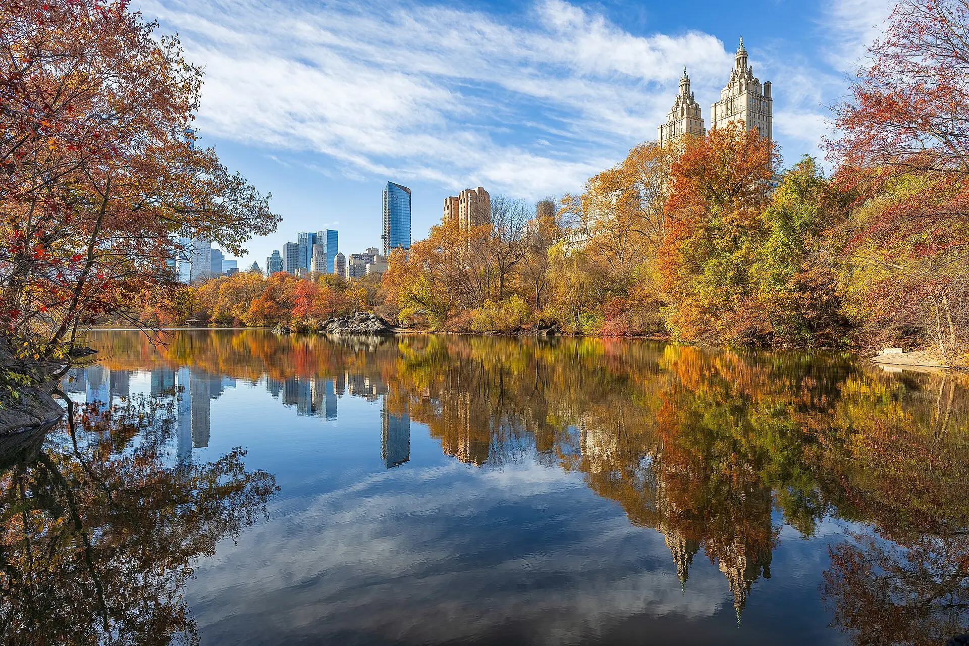 The Lake im Central Park im Herbst: rotes und gelbes Laub spiegelt sich im Wasser, rechts die Doppeltürme des San Remo