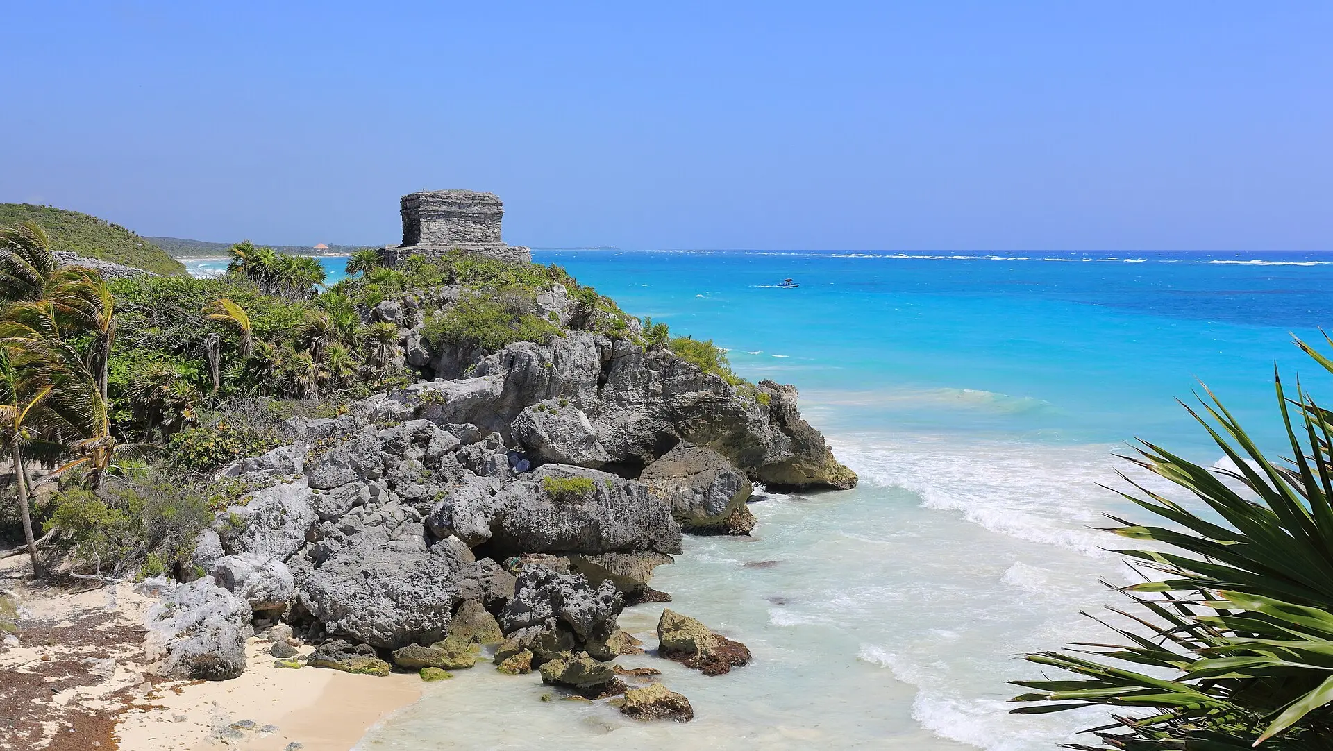 Kleiner Tempel des Windgottes auf einer Felsklippe in Tulum über türkisfarbenem Meer, davor Sandstrand mit Algenresten