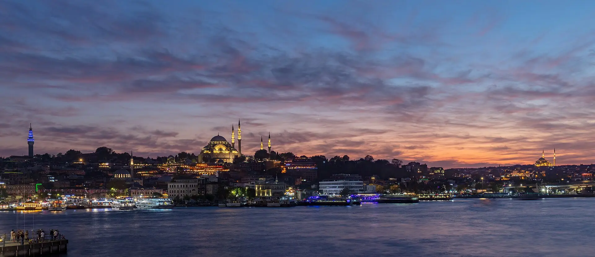 Blick über das Goldene Horn in der Abenddämmerung auf die beleuchtete Süleymaniye-Moschee, davor Anleger und Ausflugsschiffe