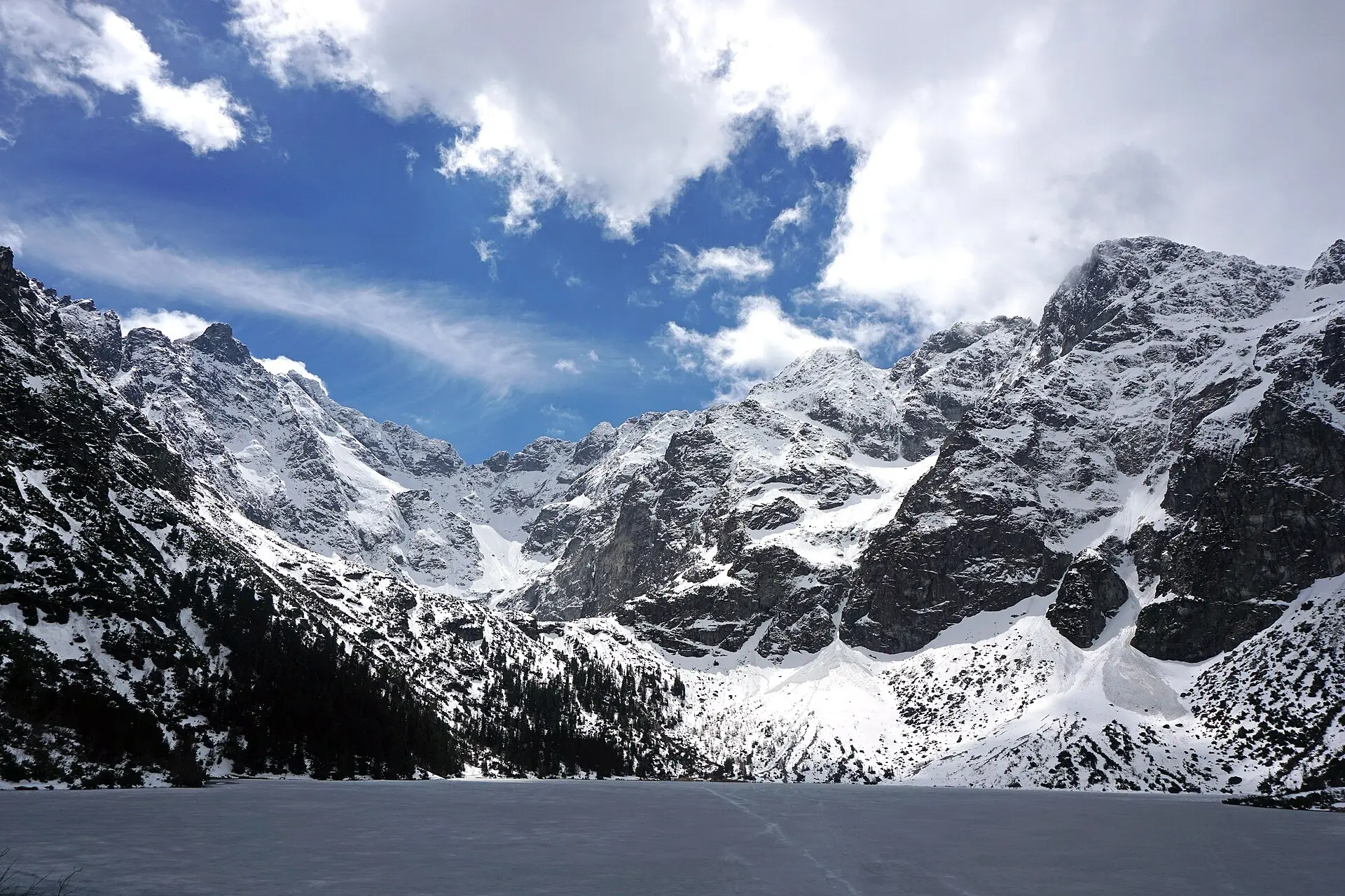 Zugefrorener, verschneiter See Morskie Oko in der Tatra im Mai, dahinter schneebedeckte Felsgipfel, links dunkler Nadelwald