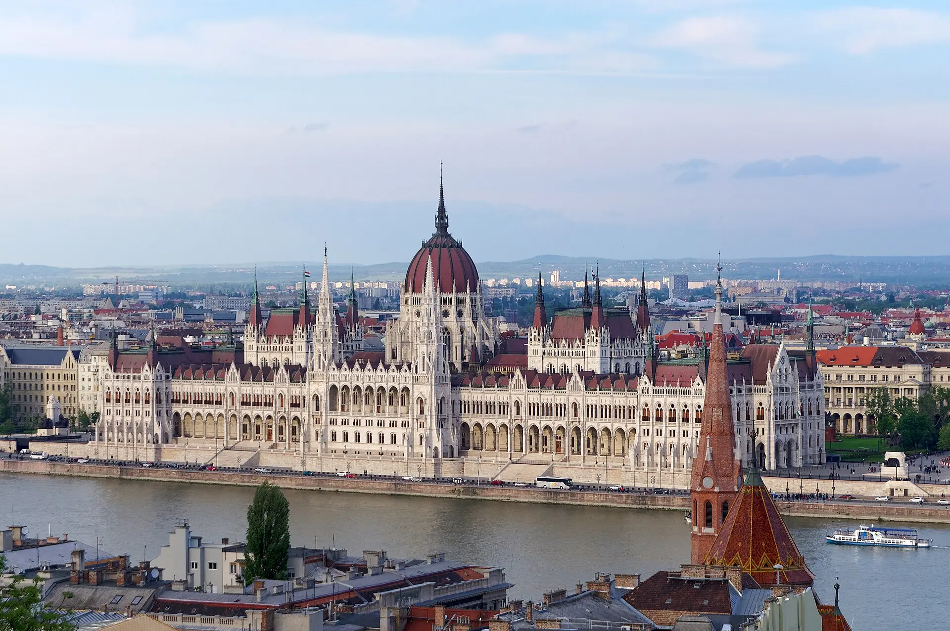 Blick über Dächer von Buda auf das Parlament mit roter Kuppel am anderen Donauufer, auf dem Fluss ein weißes Ausflugsschiff