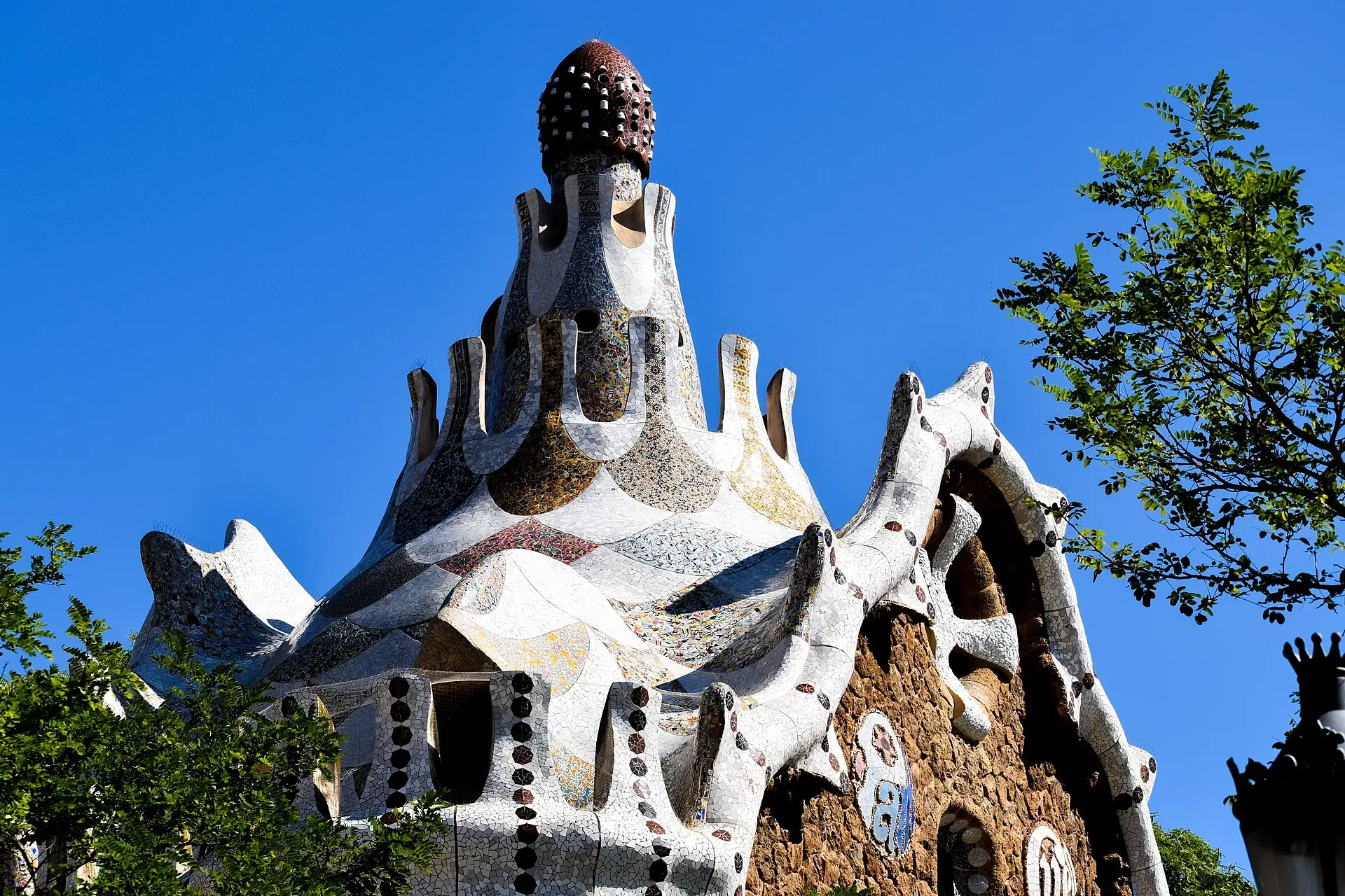 Pförtnerhaus am Eingang des Park Güell mit Mosaikturm und geschwungenem Dach aus weißer Keramik vor tiefblauem Himmel