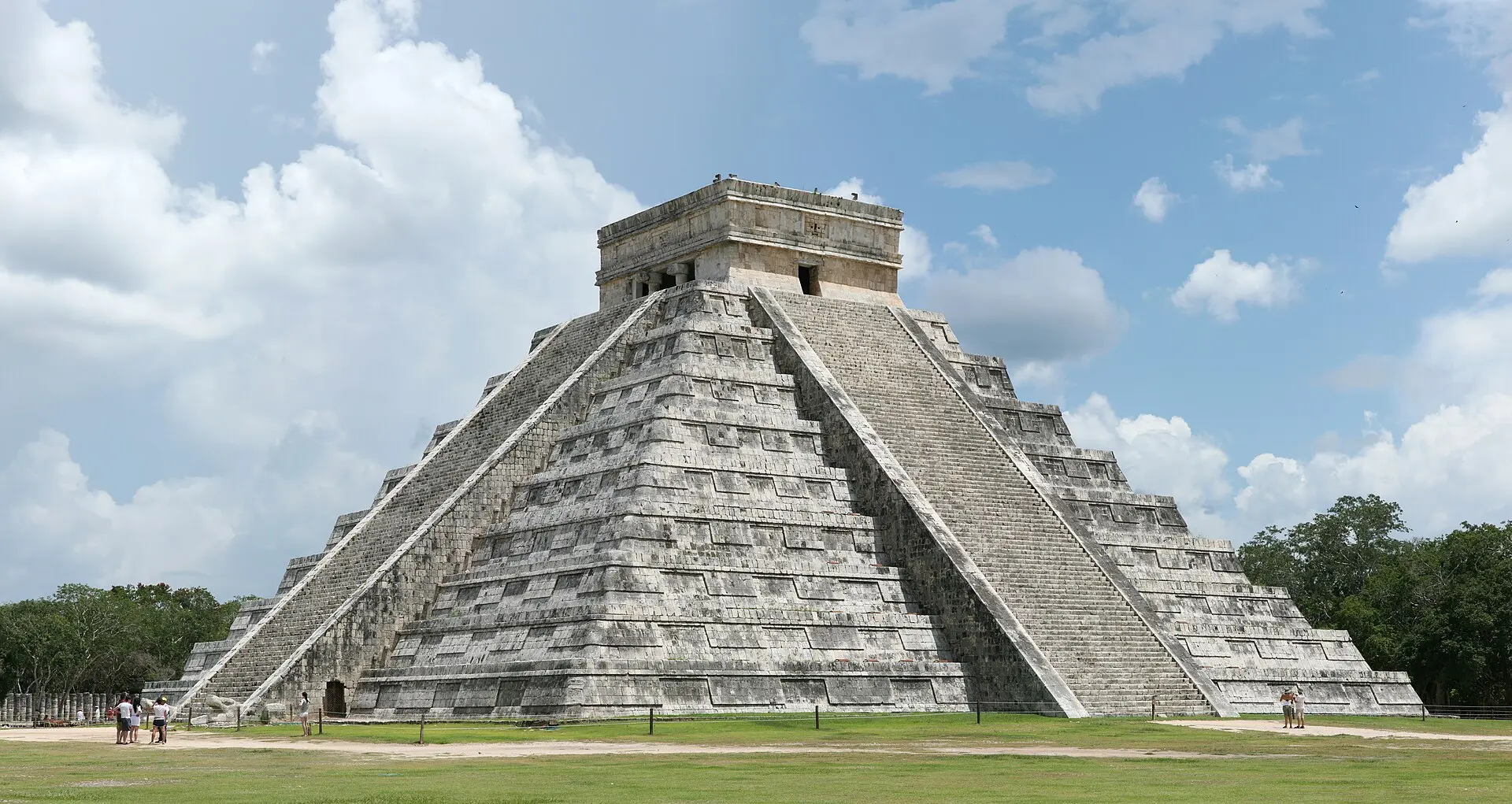 Stufenpyramide El Castillo in Chichén Itzá mit zwei Treppen und Tempel oben, davor Rasen, darüber blauer Himmel mit Wolken