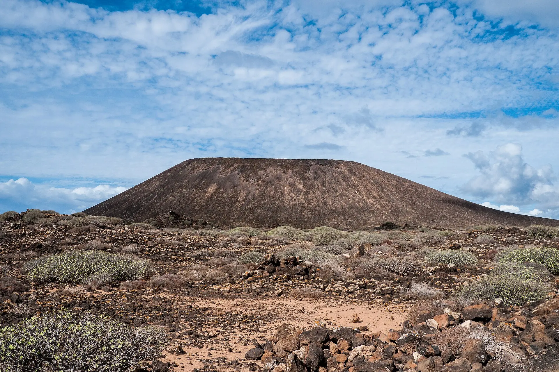 Flacher Vulkankegel La Caldera auf der Isla de Lobos hinter einer steinigen Ebene mit graugrünen Sträuchern, darüber Schäfchenwolken