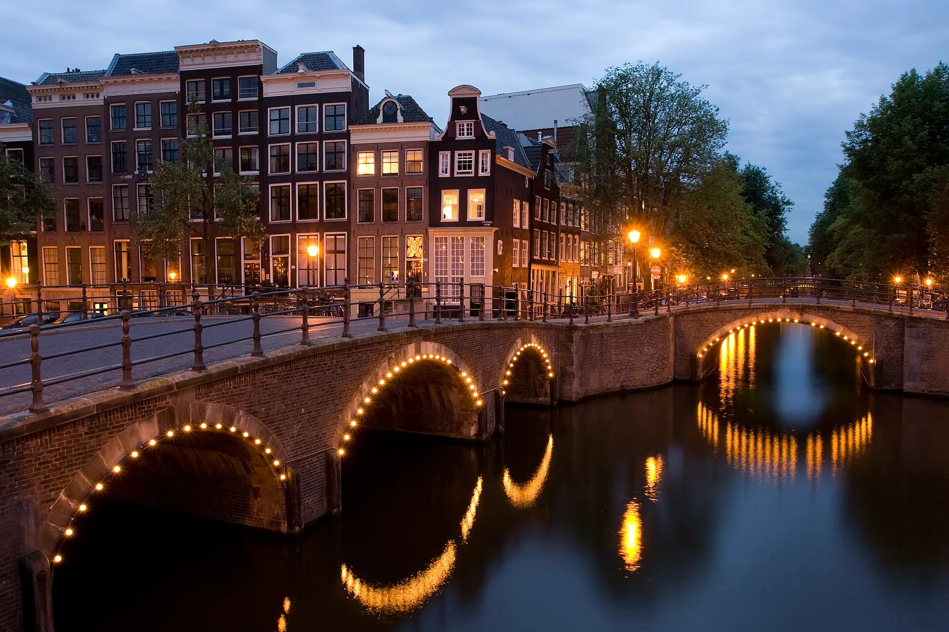 Zwei gemauerte Bogenbrücken mit Lichterketten an der Kreuzung von Keizersgracht und Reguliersgracht in der Abenddämmerung
