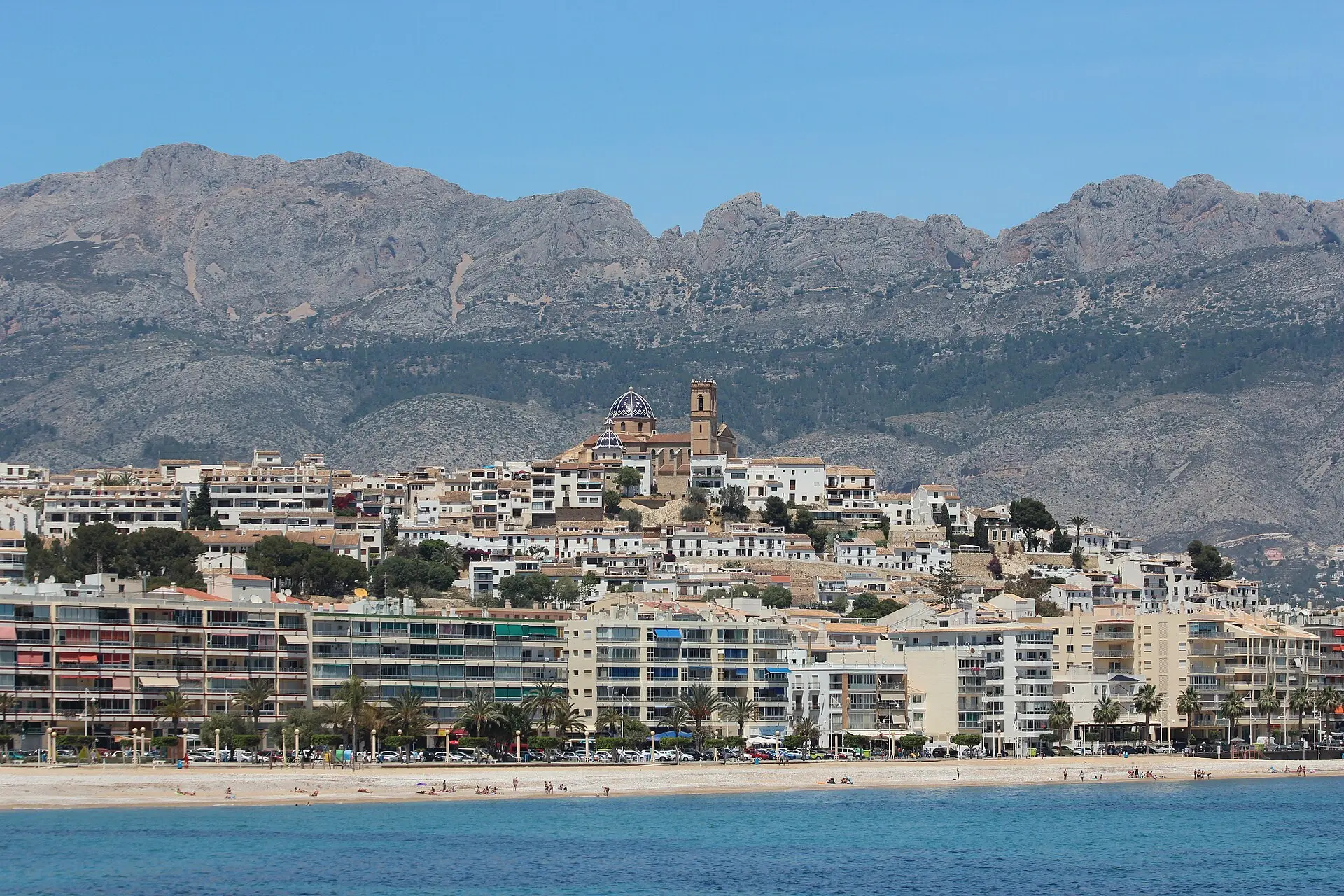 Altea vom Hafen aus: Altstadt mit blau-weißer Kirchenkuppel und Glockenturm, darunter Apartmenthäuser, ein schmaler Strand und Berge