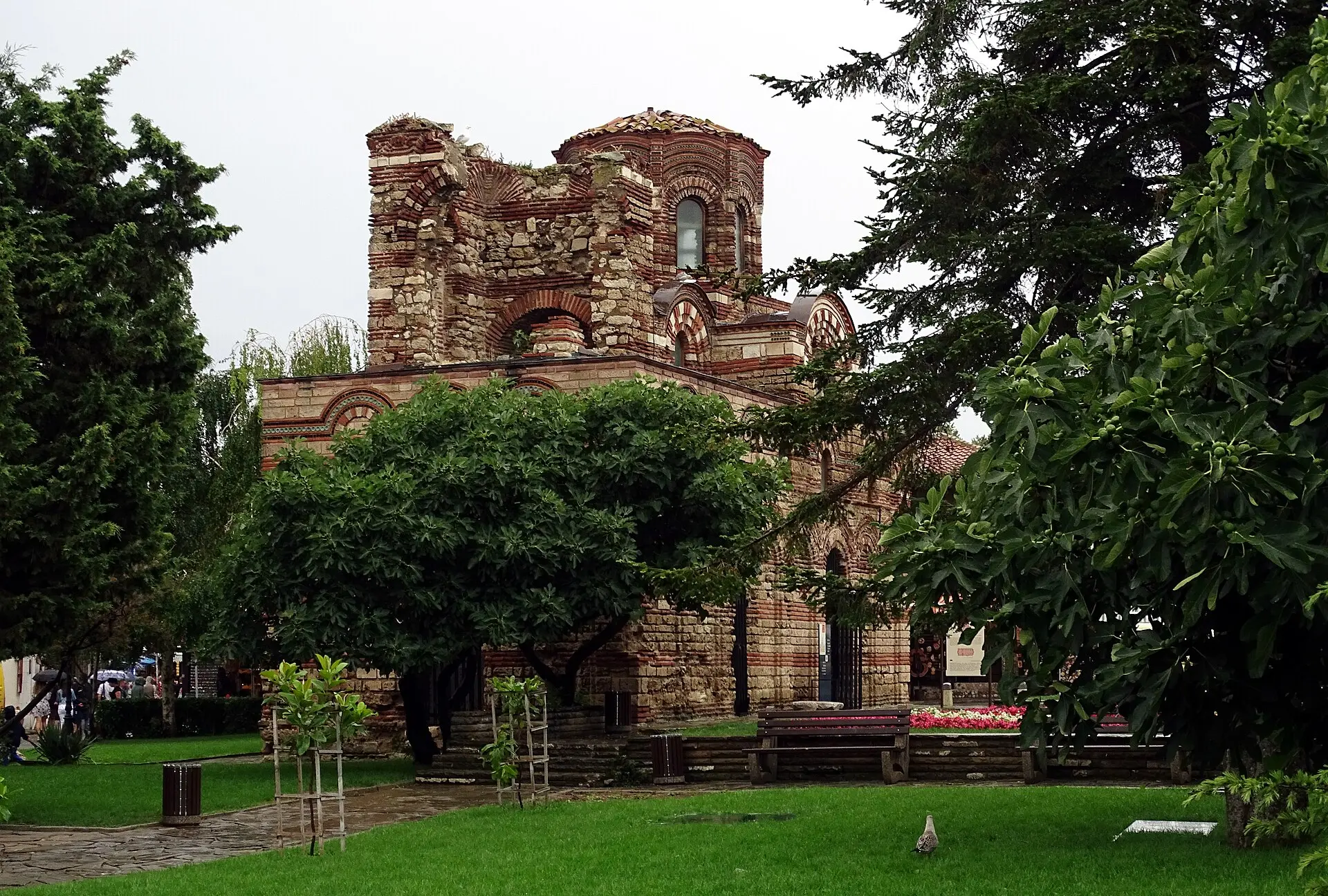 Kirche Christus Pantokrator in Nessebar aus Ziegel- und Steinbändern mit Zierbögen, halb hinter Bäumen, davor Rasen und eine Bank