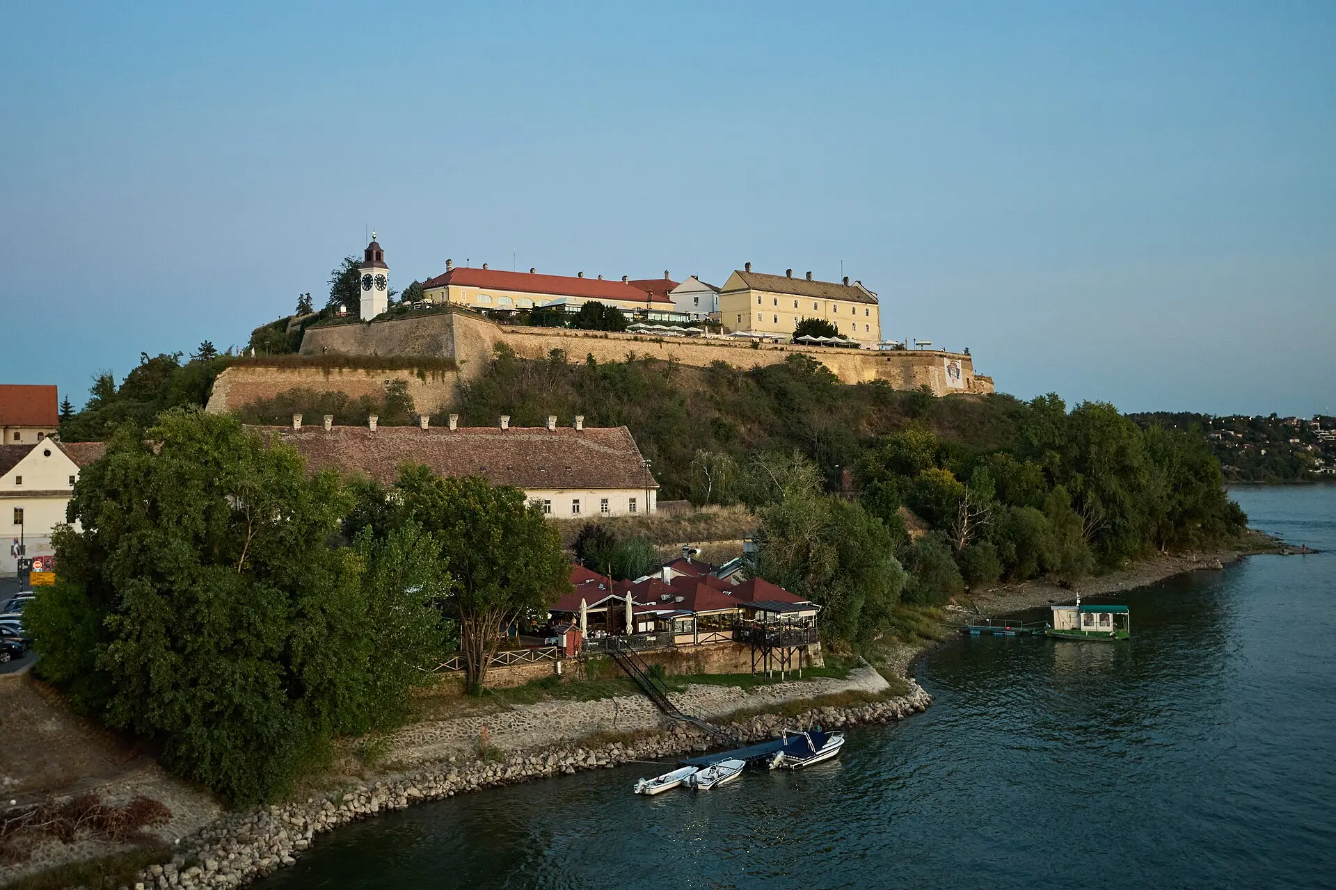 Festung Petrovaradin auf einem grünen Hügel im Abendlicht, links der weiße Uhrturm, unten ein Lokal am Ufer und kleine Boote