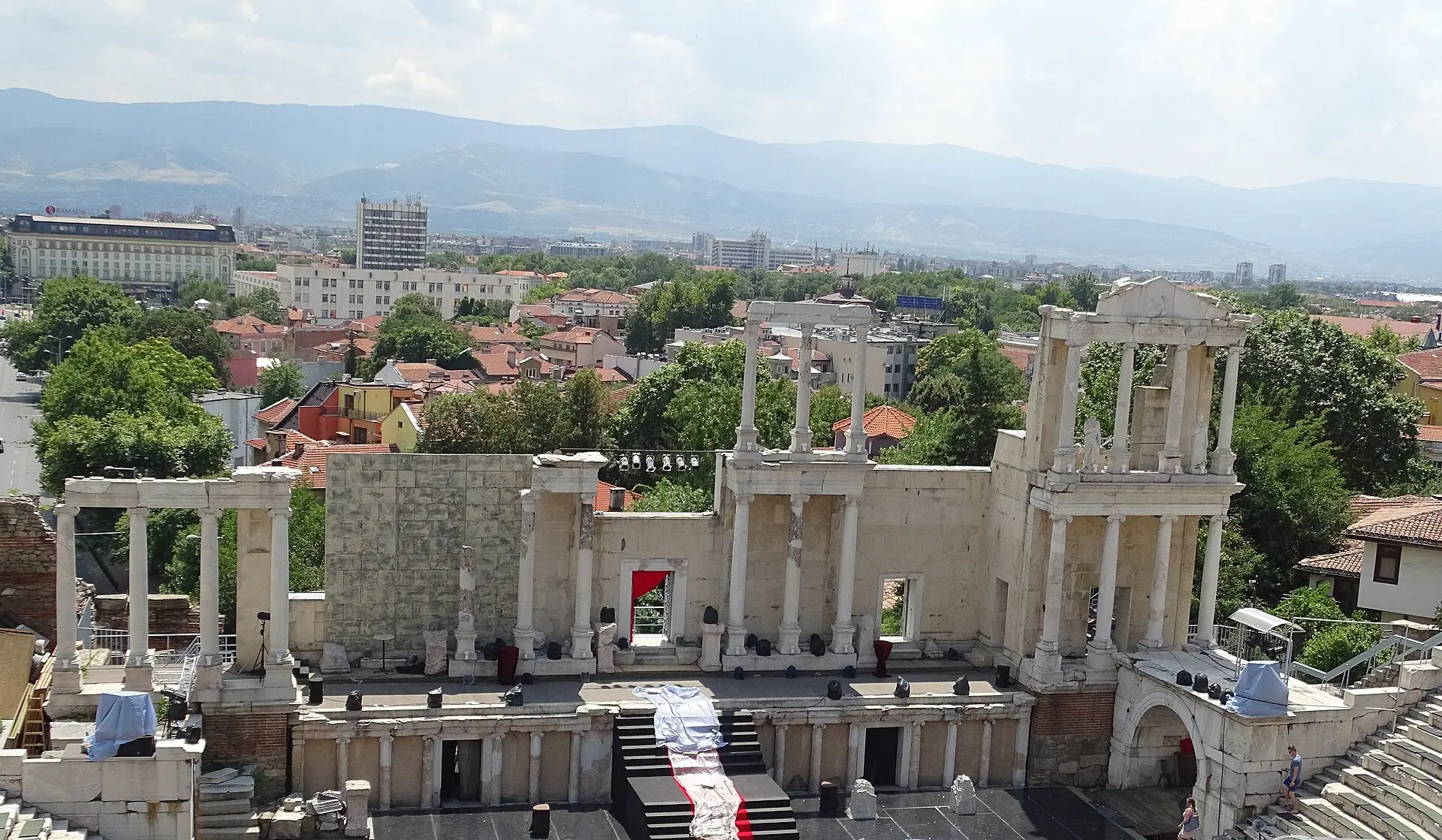 Antikes Theater von Plovdiv von oben: Bühnenwand mit weißen Säulen und Scheinwerfern, dahinter Dächer der Stadt und Berge im Dunst