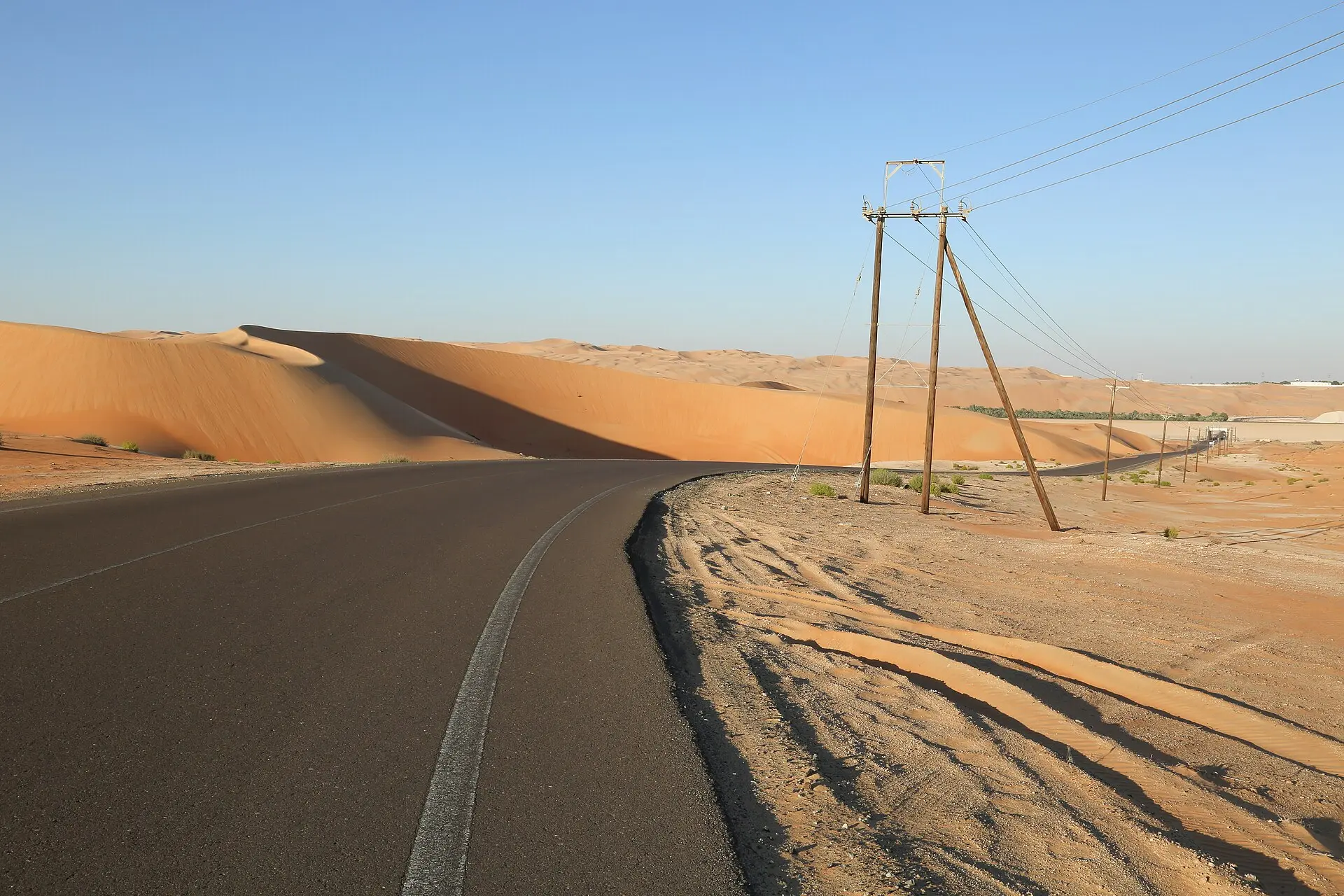 Asphaltstraße an hohen orangefarbenen Sanddünen der Liwa-Wüste vorbei, rechts hölzerne Strommasten, wolkenloser blauer Himmel