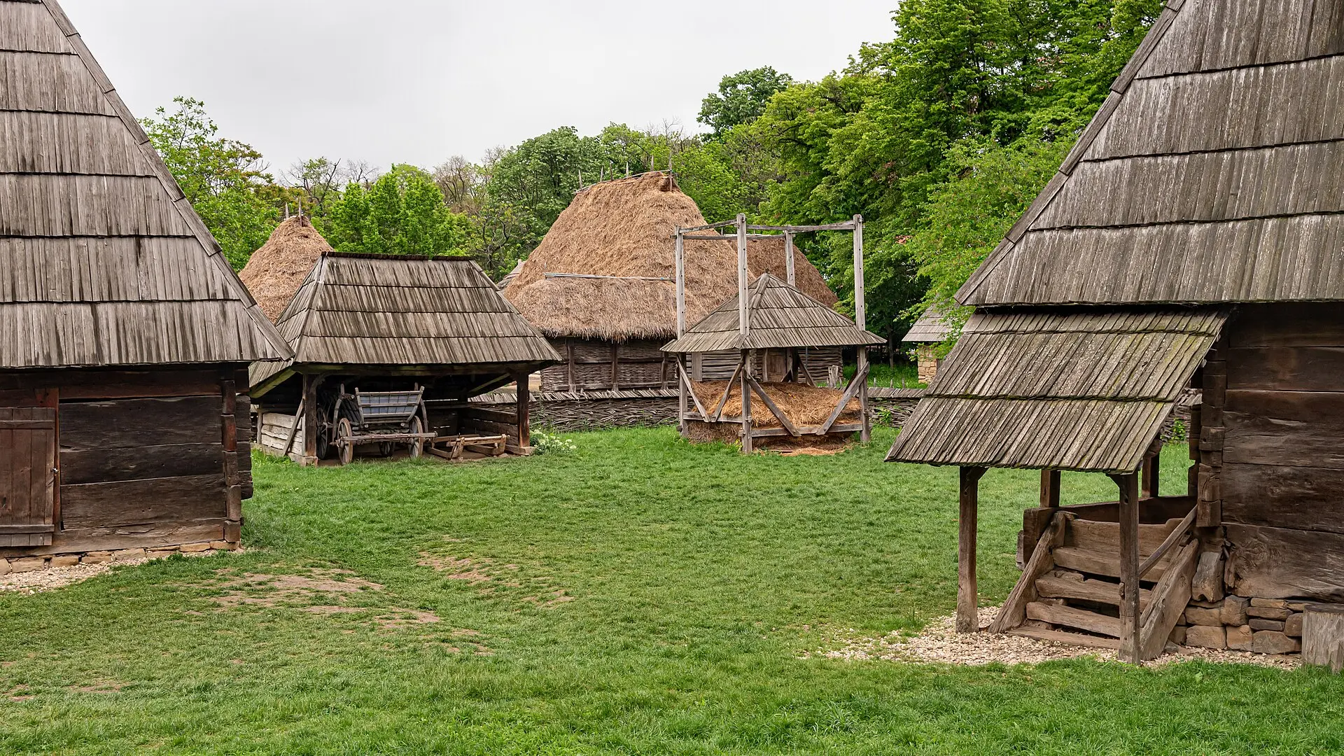 Holzhäuser und Scheunen mit Schindel- und Strohdächern um eine Wiese im Dorfmuseum, ein Holzwagen unter einem Vordach
