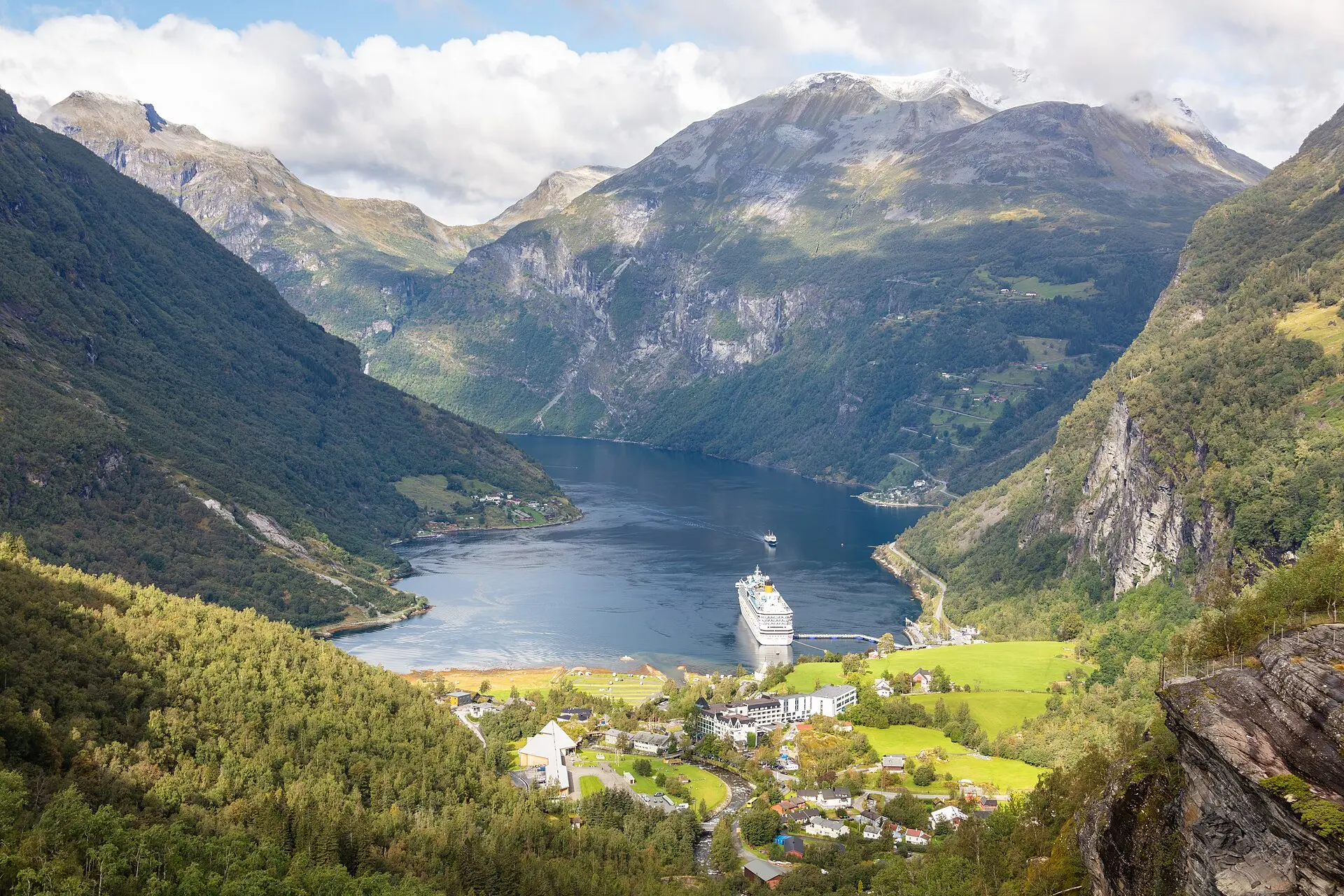 Blick von Flydalsjuvet auf den Geirangerfjord mit steilen grünen Hängen, einem Kreuzfahrtschiff und dem Dorf Geiranger