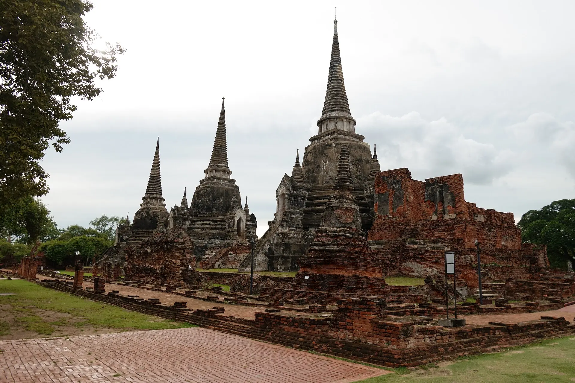 Drei glockenförmige Chedis des Wat Phra Si Sanphet unter bewölktem Himmel, rechts Ziegelruinen, vorn Backsteinfundamente und ein Weg