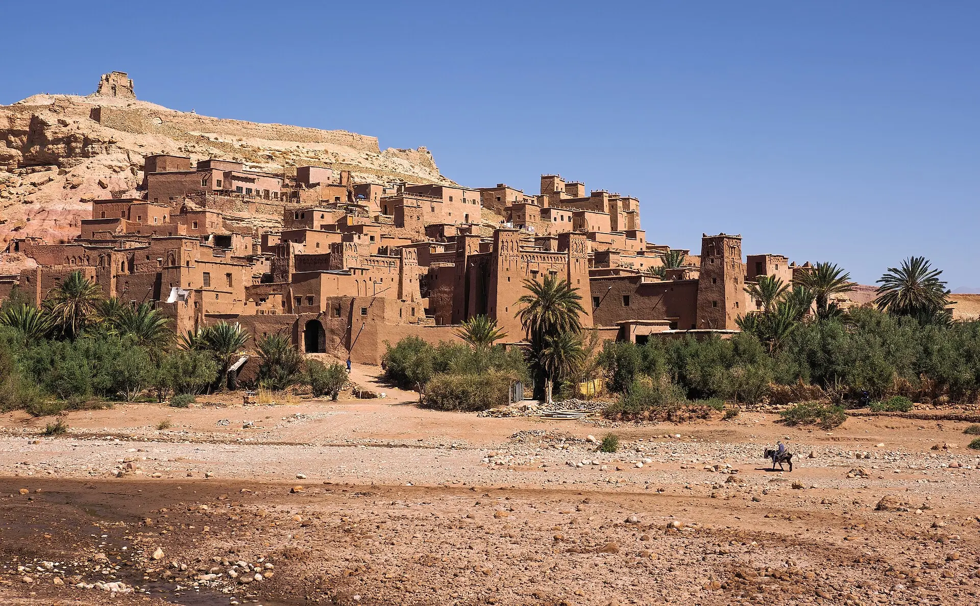 Lehmbauten des Ksar Aït-Ben-Haddou am Hang unter einer Speicherburg, davor Palmen und ein trockenes, steiniges Flussbett