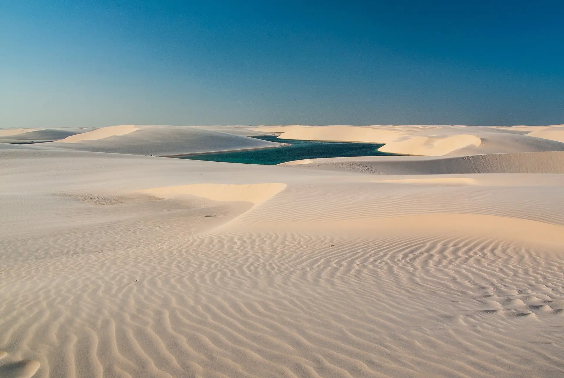 Weiße Dünen mit Rippelmarken, dahinter eine blaugrüne Lagune zwischen Dünenkämmen unter wolkenlosem blauem Himmel
