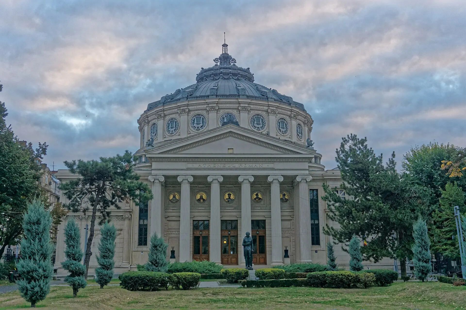 Das Rumänische Athenäum im Morgenlicht: Säulenportal mit Inschrift, Kuppel, davor eine Statue, Rasen, Hecken und Nadelbäume