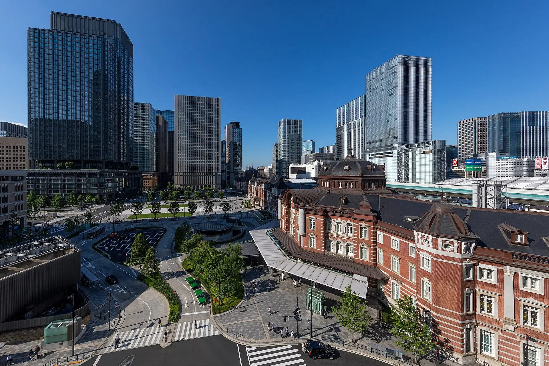 Rotes Backsteingebäude des Bahnhofs Tokio mit Kuppel, davor der Bahnhofsplatz mit Grünflächen und Taxis, dahinter Glashochhäuser