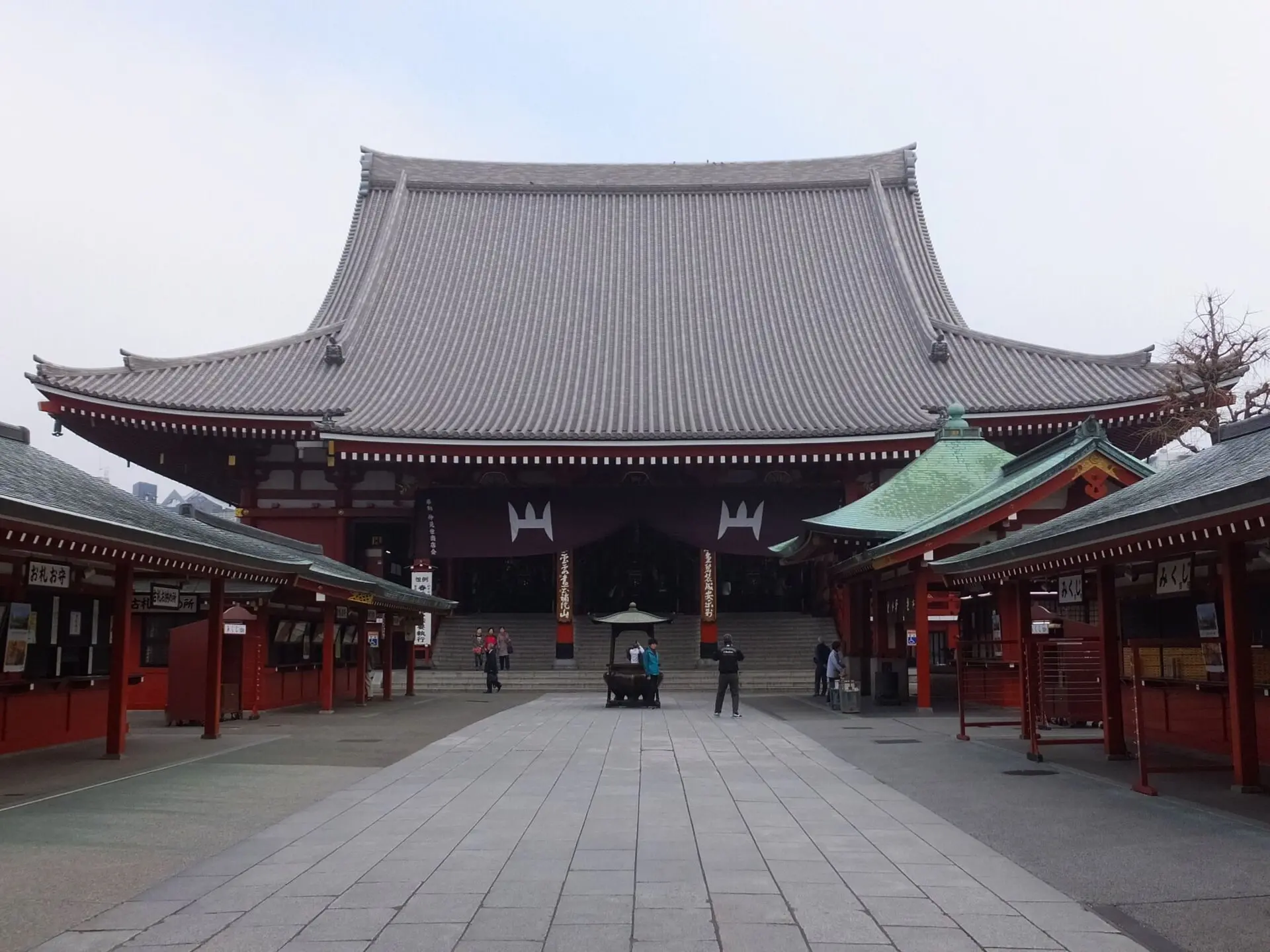 Haupthalle des Senso-ji mit steilem grauem Ziegeldach, davor ein Plattenweg mit Räucherbecken und rote Nebengebäude