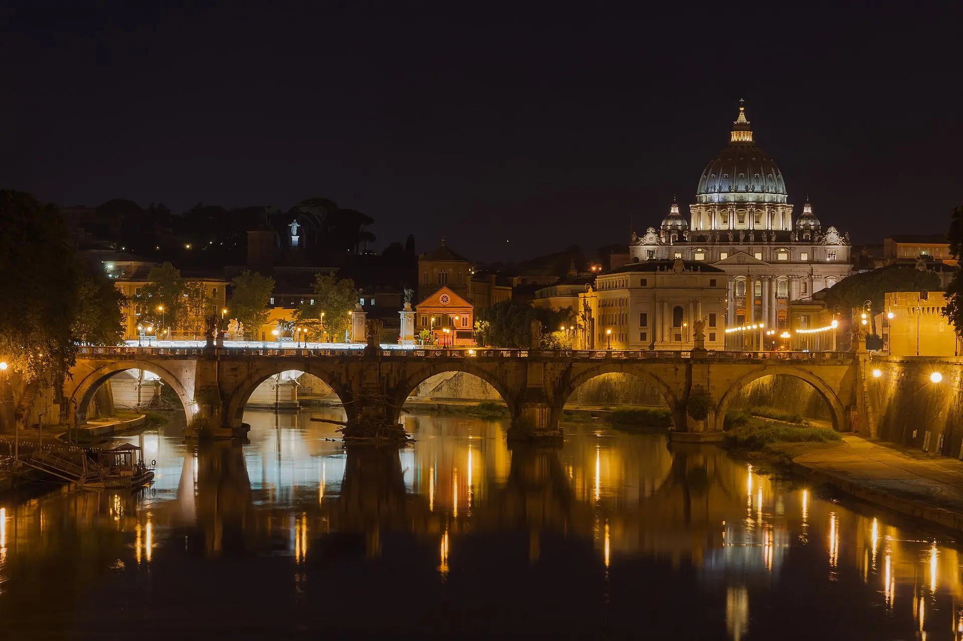 Engelsbrücke über dem Tiber bei Nacht, dahinter die beleuchtete Kuppel des Petersdoms, die Lichter spiegeln sich im Wasser