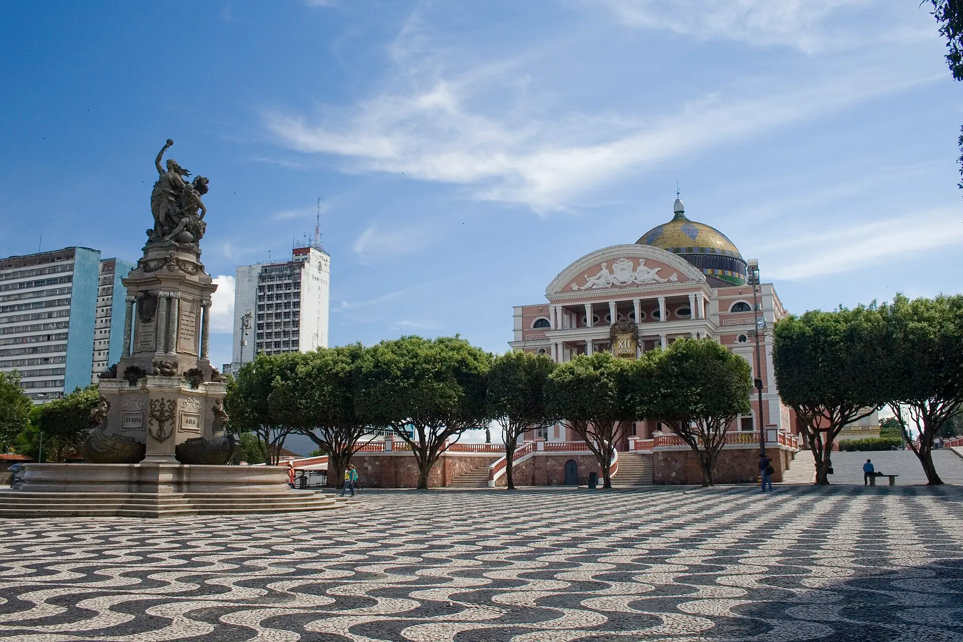 Rosa Fassade des Teatro Amazonas mit Säulenportal und bunt gemusterter Kuppel, davor ein Platz mit schwarz-weißem Wellenpflaster